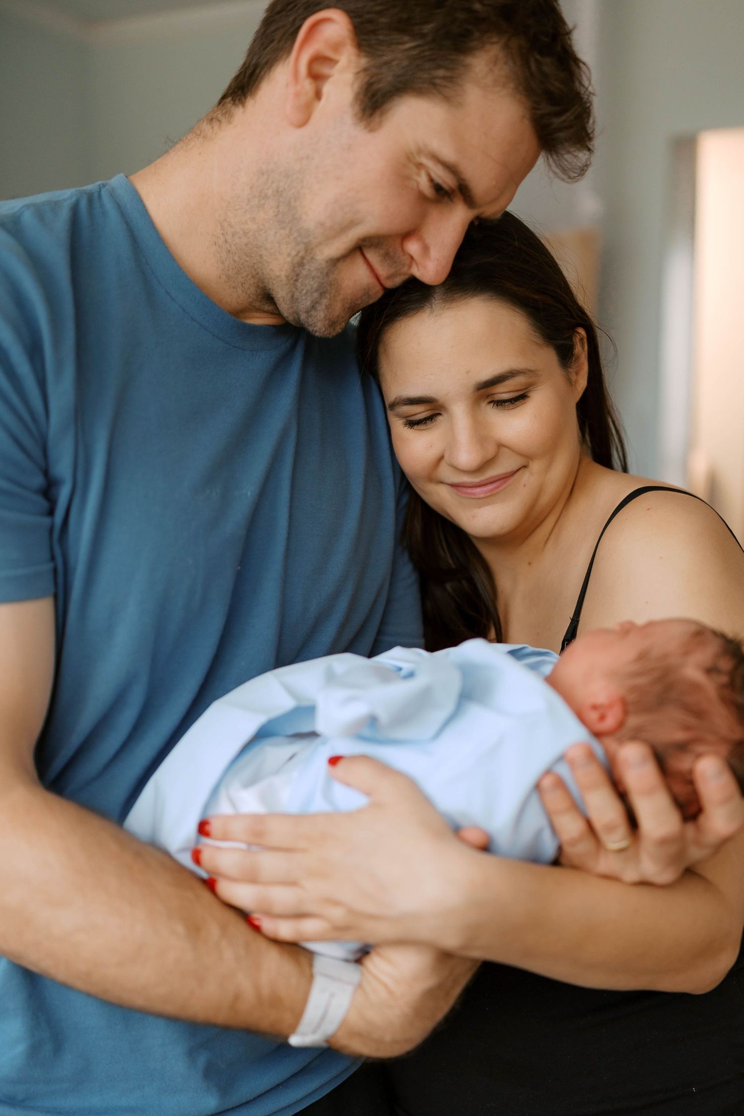 New parents smiling lovingly at their swaddled newborn baby wrapped in a blue blanket indoors.