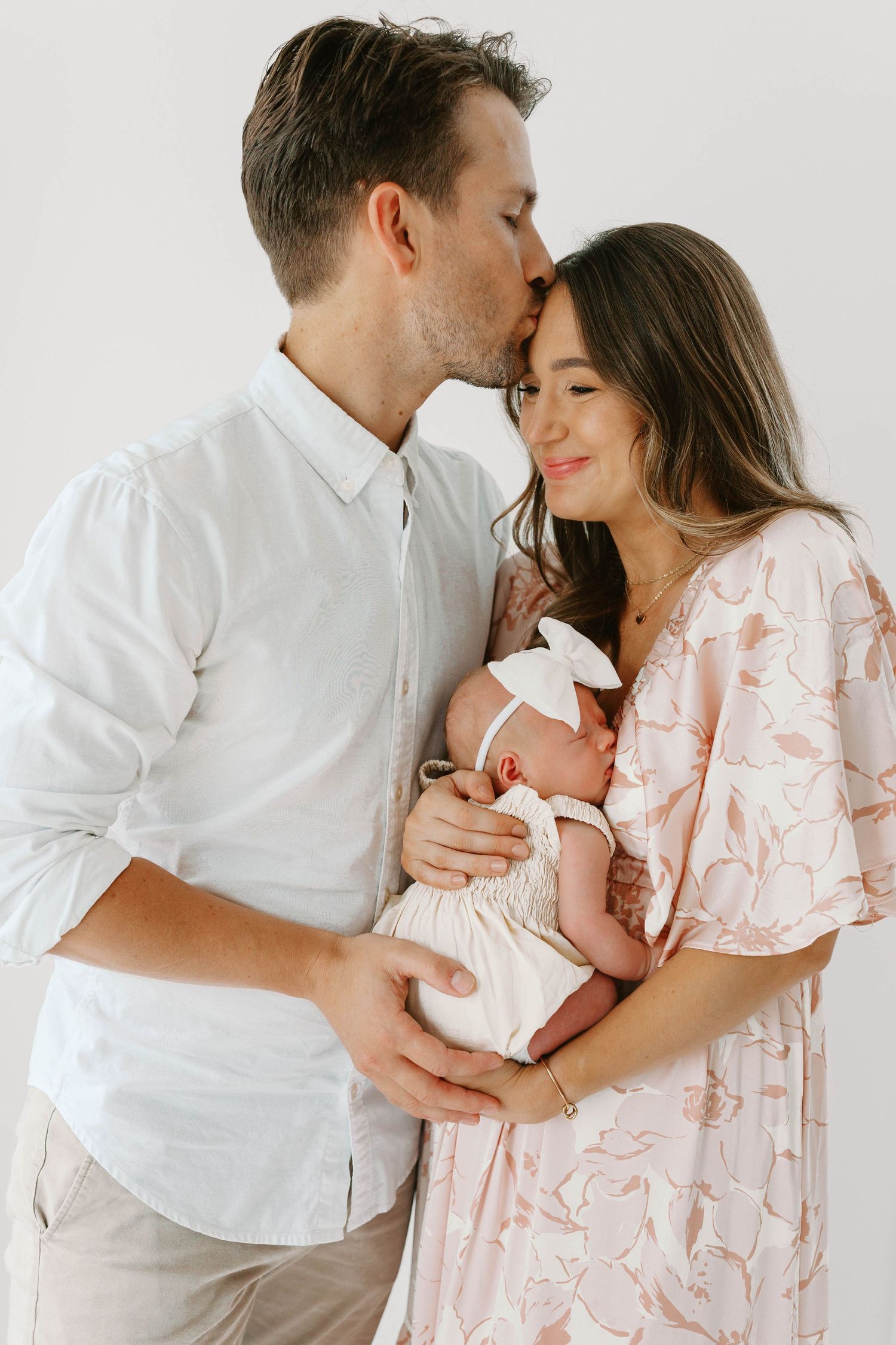 A couple tenderly holds their newborn baby, the man kissing the woman's forehead, in a bright studio setting in Kirkwood, MO.