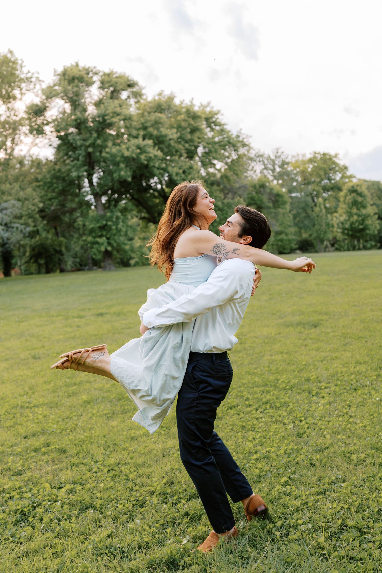 Man joyfully lifts a laughing woman in a blue dress while spinning in Lafayette square park.