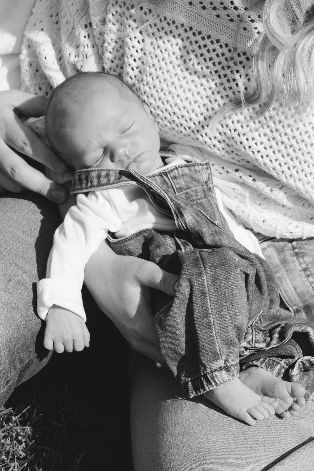 Black and white photo of sleeping newborn in denim overalls cradled in mother's arms outdoors.