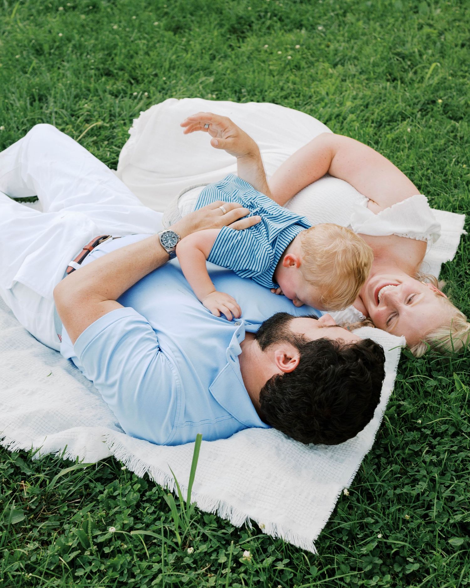 Family of three lying on white blanket in grass, laughing and bonding with baby.
