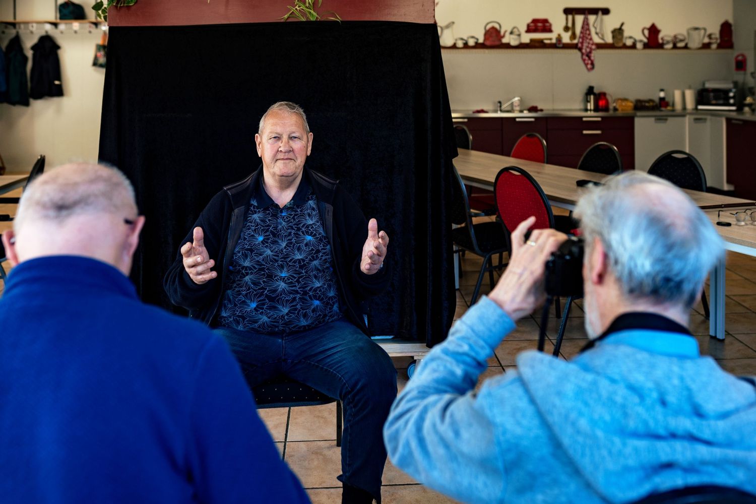 A photography session in progress with someone in a blue patterned shirt being photographed against a black backdrop.