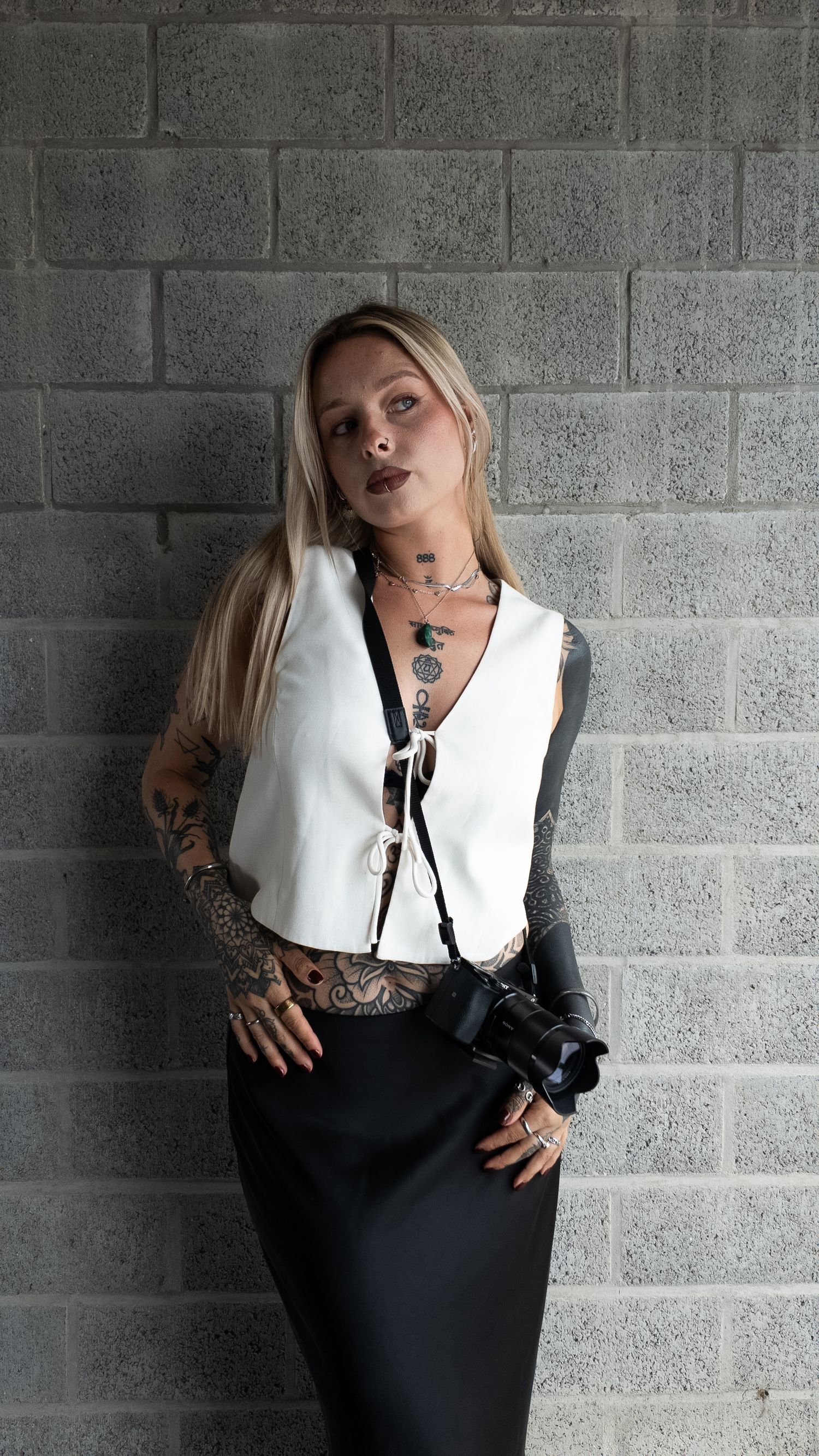Tattooed blonde woman in white vest and black skirt holding camera against grey brick wall. By Soft Sacred Studios.