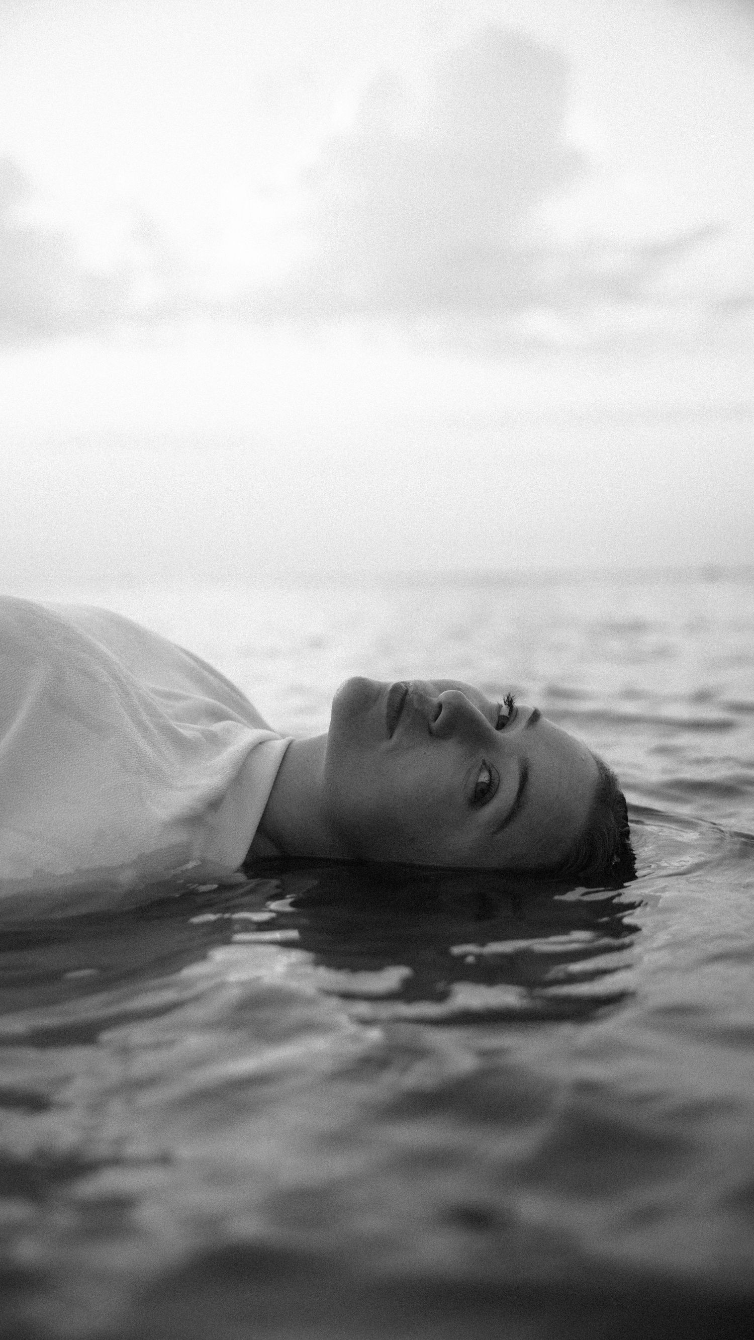 Black and white portrait of a young woman with wet hair lying in the sea. By Soft Sacred Studios.
