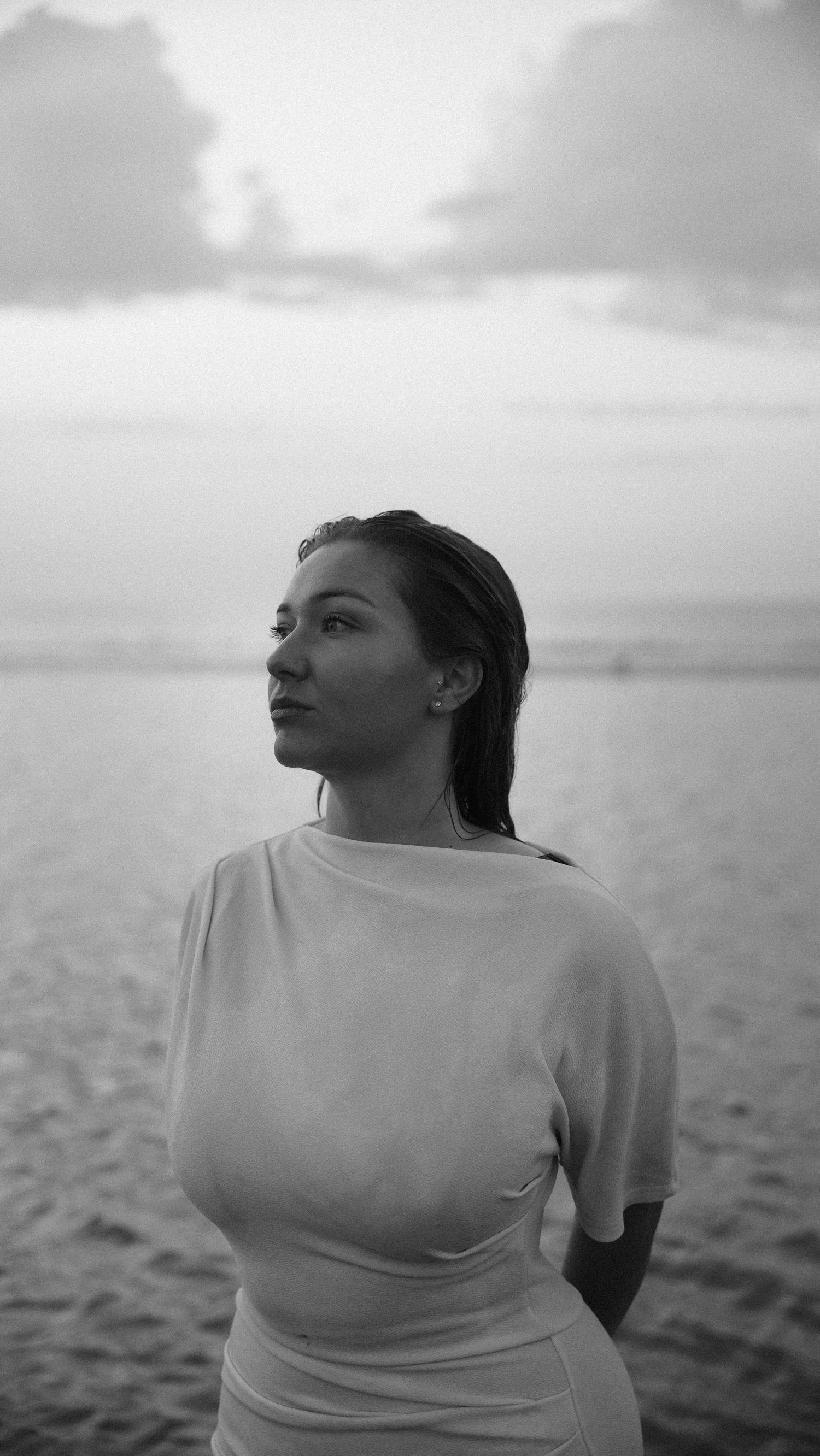 Black and white photo of a woman in white dress standing by the calm ocean at dusk. by Soft Sacred Studios