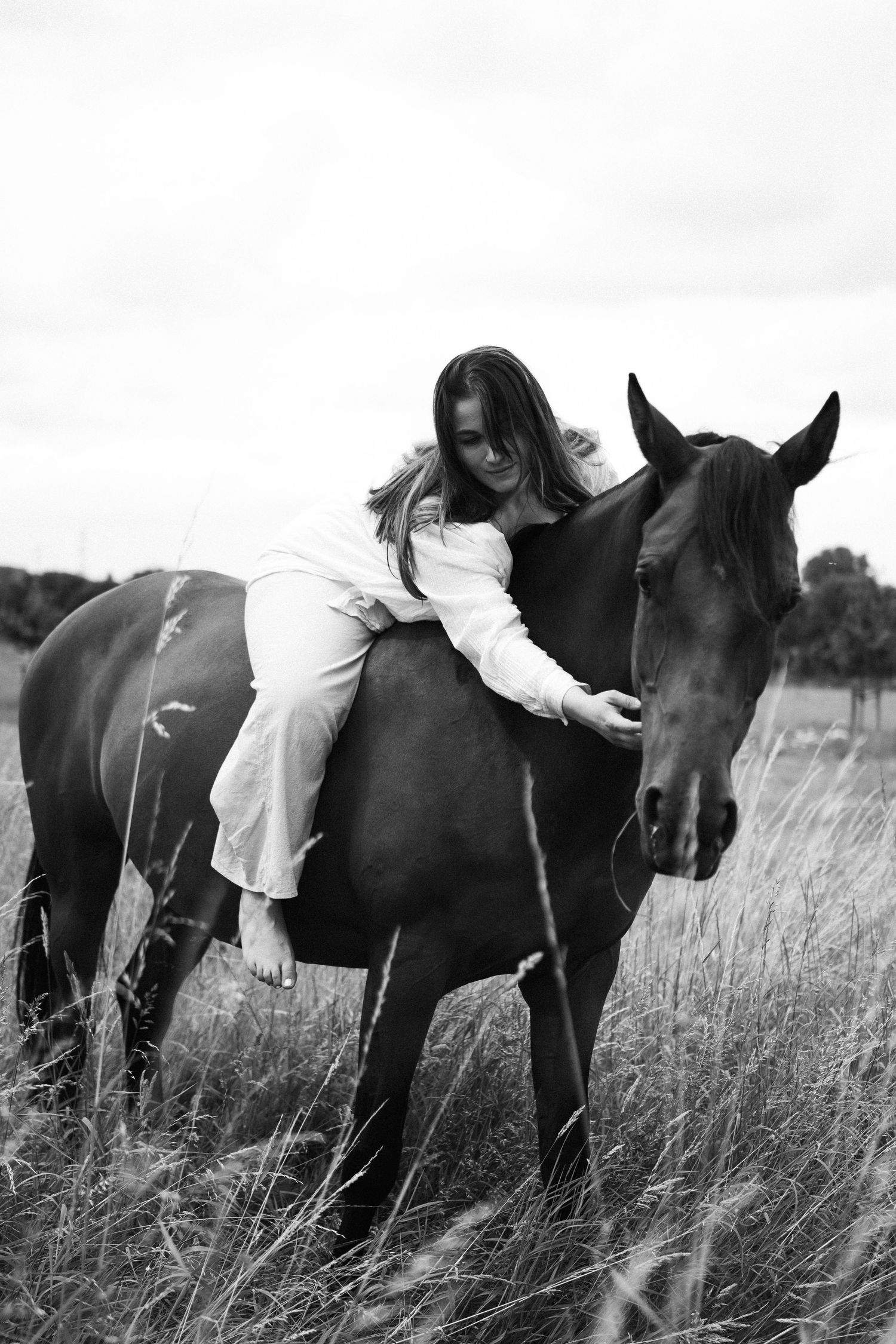 Black and white photo of woman lying on horseback embracing a horse in a field. By Soft Sacred Studios.
