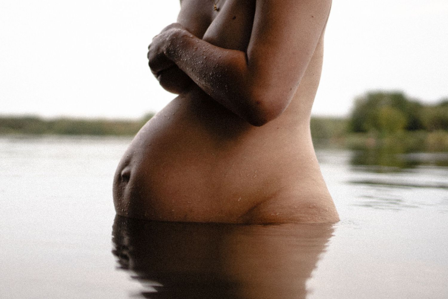 Warm-toned artistic maternity photo of pregnant woman standing in natural lake water. By Soft Sacred Studios.