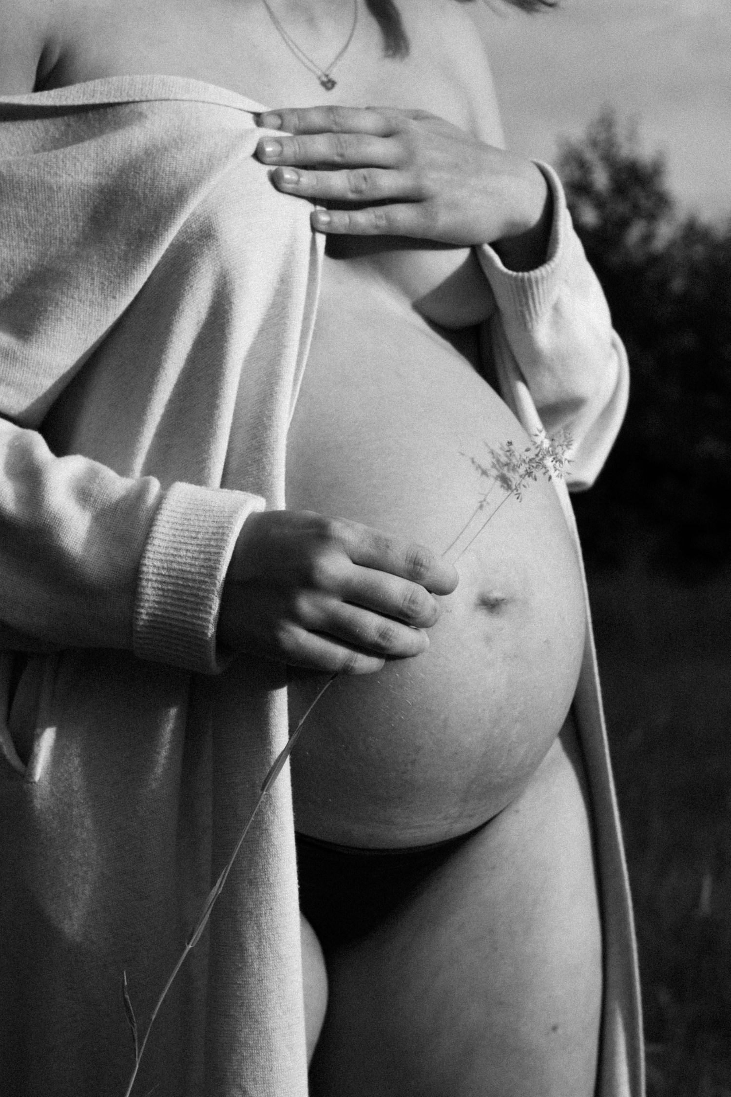 Black and white close-up of pregnant woman holding wildflower against bare baby bump wearing open cardigan. by Soft Sacred Studios