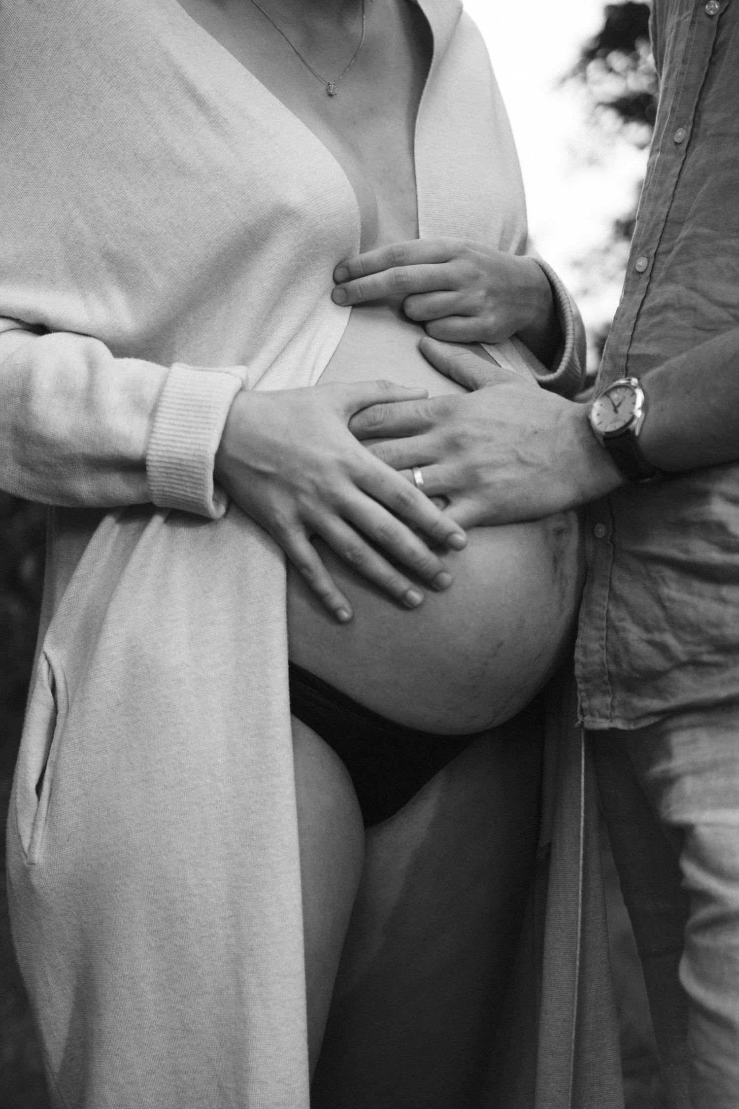 Black and white close-up of couple tenderly holding pregnant belly outdoors during maternity shoot. By Soft Sacred Studios.