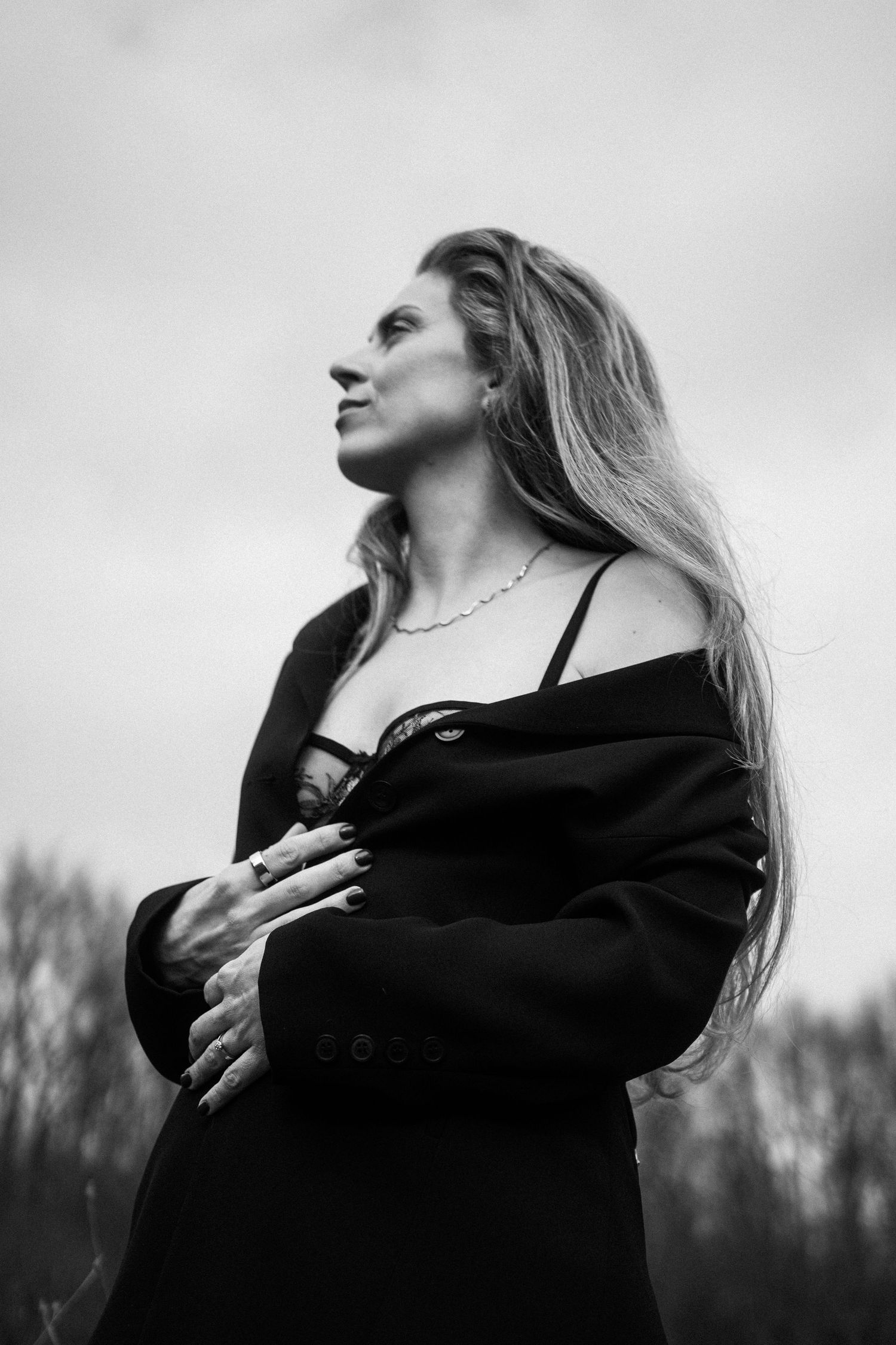 Black and white portrait of long-haired blonde woman in black off-shoulder coat gazing sideways outdoors. By Soft Sacred Studios.