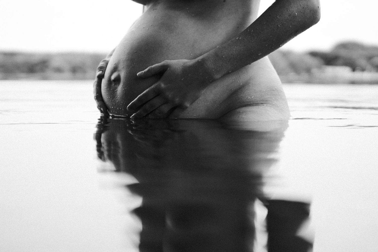 Black and white maternity photo of pregnant woman standing in water with hands cradling baby bump. By Soft Sacred Studios.