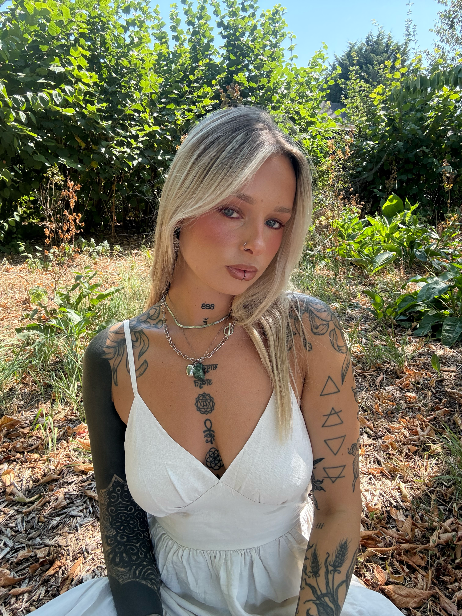 Tattooed blonde woman in white dress with layered necklaces posing in a sunny garden. By Soft Sacred Studios.