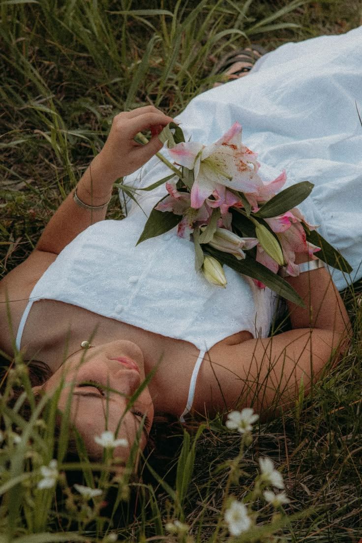 Woman lying in grass holding pink lily flowers wearing a white dress. By Soft Sacred Studios.