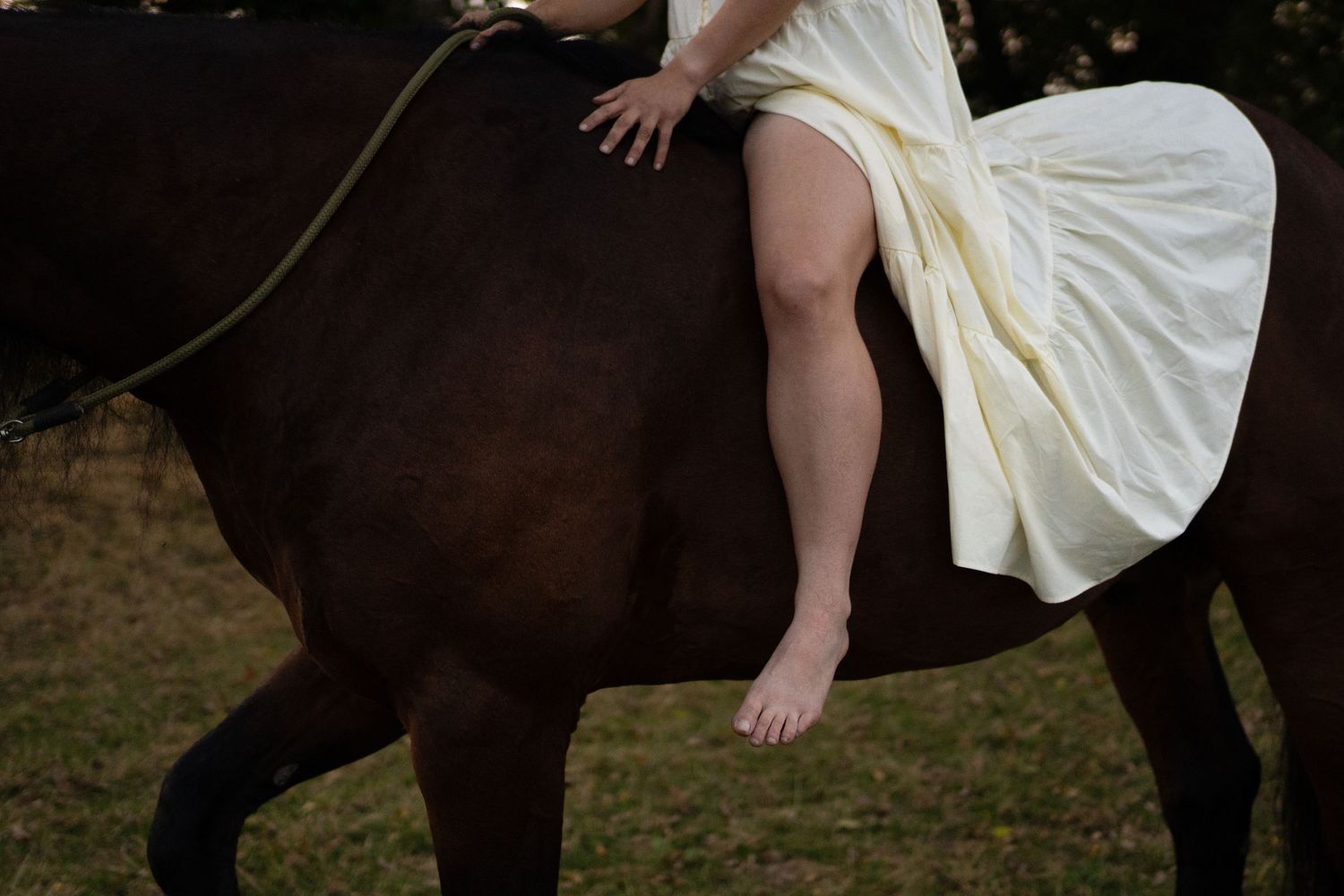 Close-up of barefoot woman in white dress sitting bareback on a dark brown horse outdoors. by Soft Sacred Studios