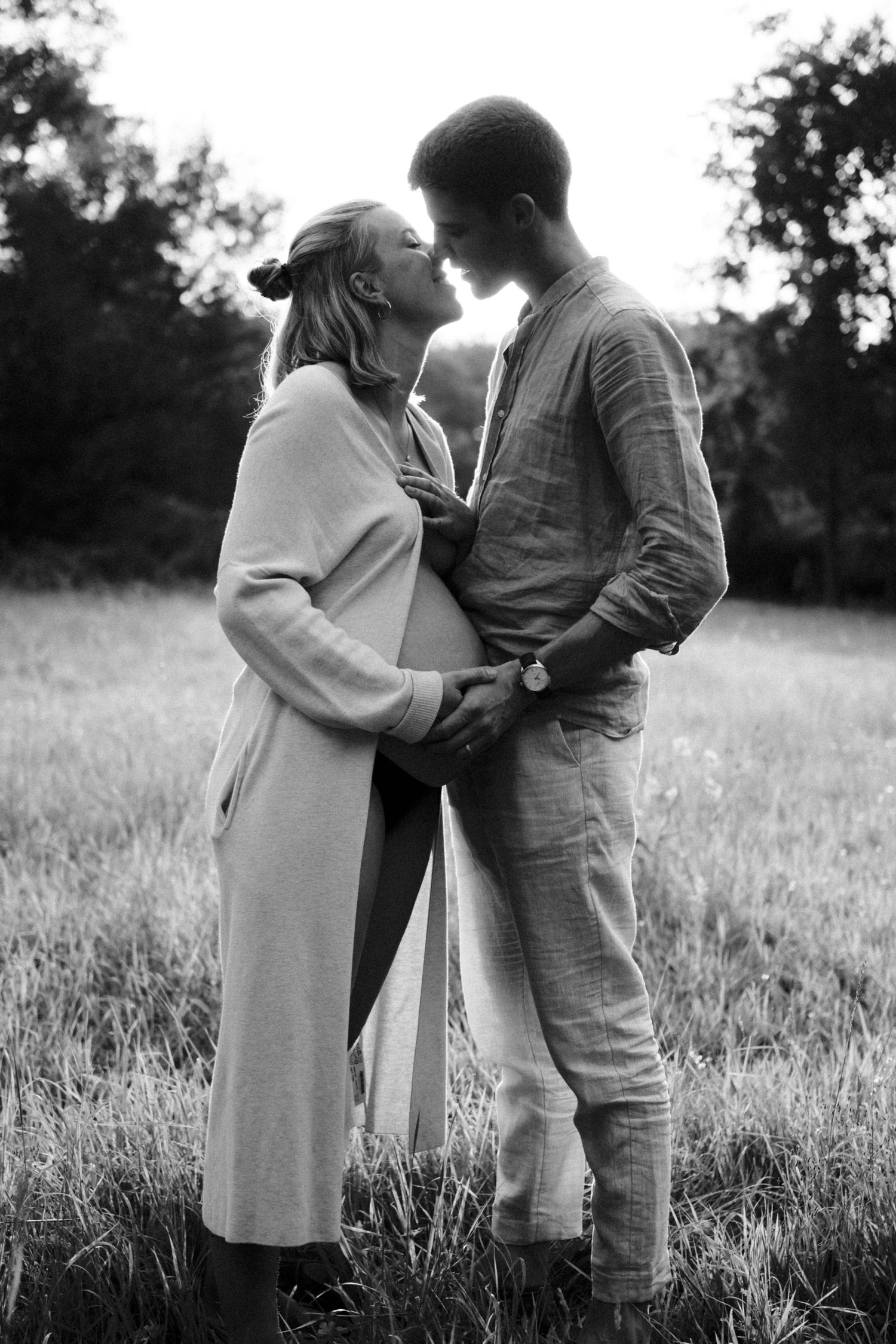 Black and white maternity photo of couple standing close together in a lush green meadow. By Soft Sacred Studios.