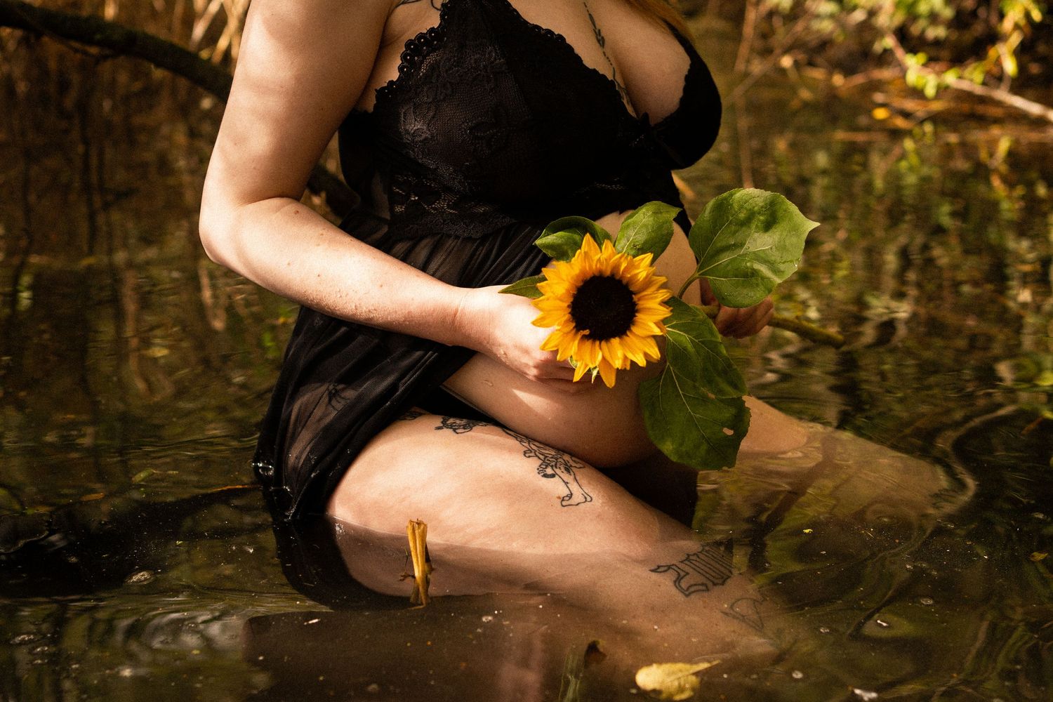 Pregnant woman in black lace dress holding vibrant sunflower while sitting in golden-lit woodland stream. By Soft Sacred Studios.
