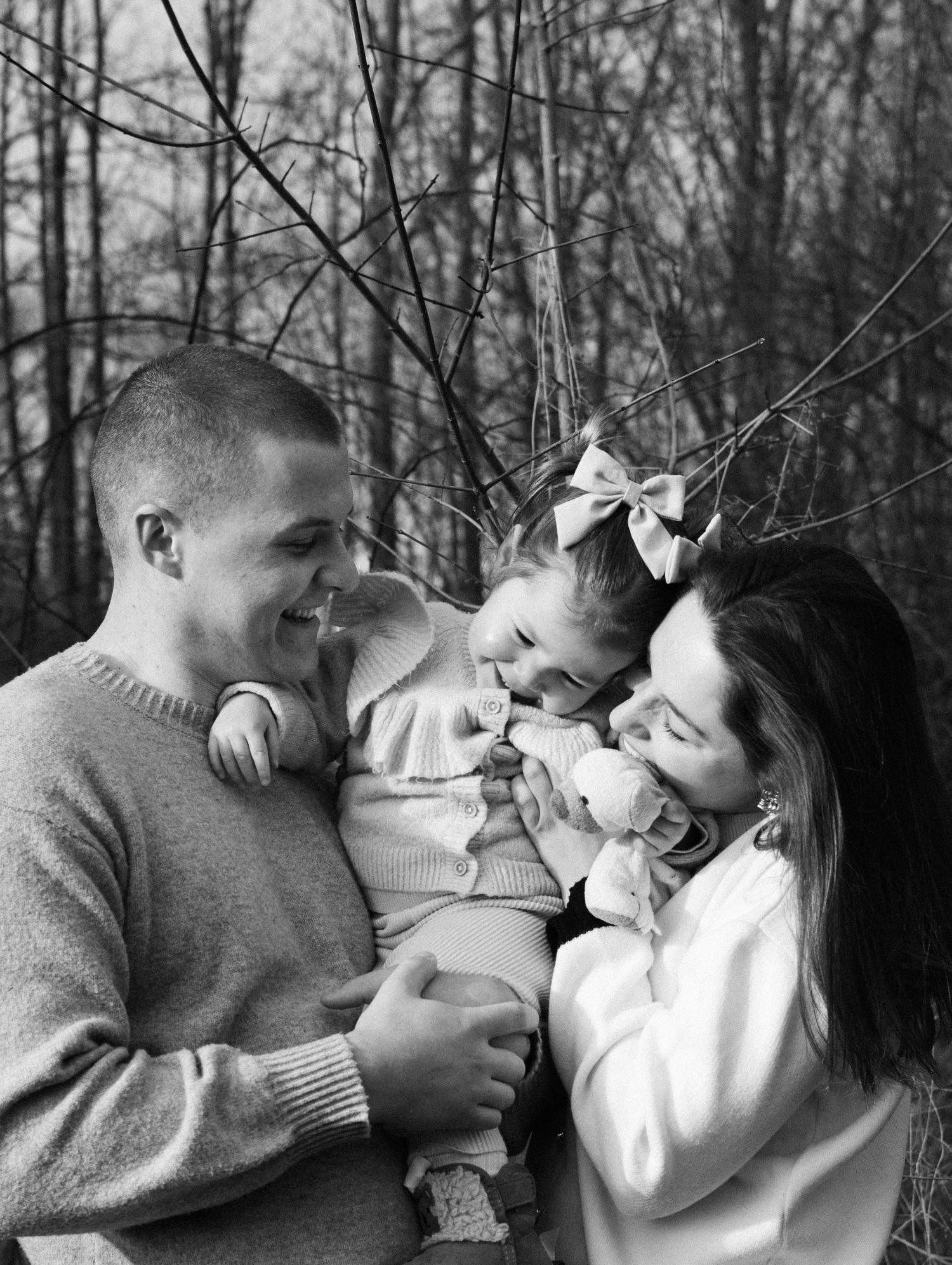 Black and white family portrait of parents smiling at laughing baby girl with bow in wooded setting. By Soft Sacred Studios.