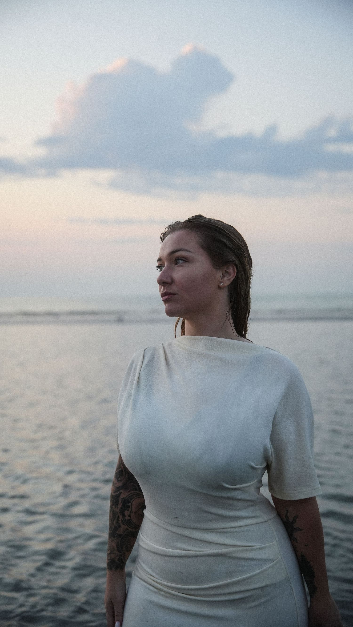 Tattooed woman in white fitted dress posing by the ocean at twilight with a contemplative gaze. by Soft Sacred Studios