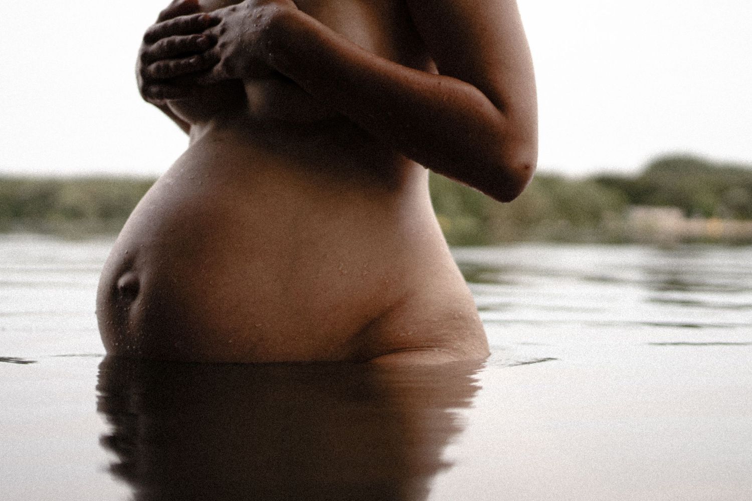 Artistic maternity photo of pregnant woman standing in water with hands clasped over her chest. By Soft Sacred Studios.