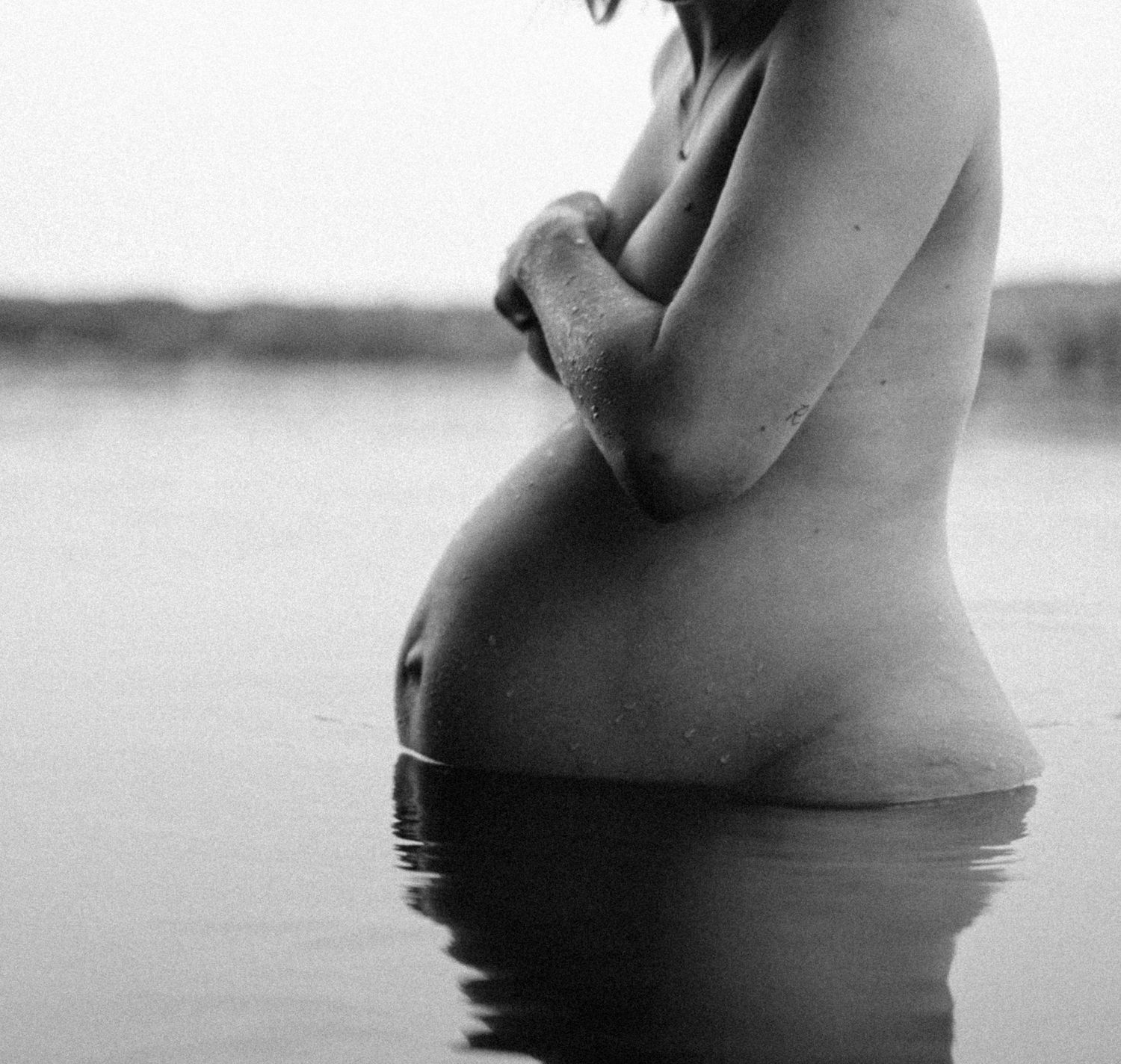 Black and white film photo of nude pregnant woman standing in lake water cradling her belly. By Soft Sacred Studios.