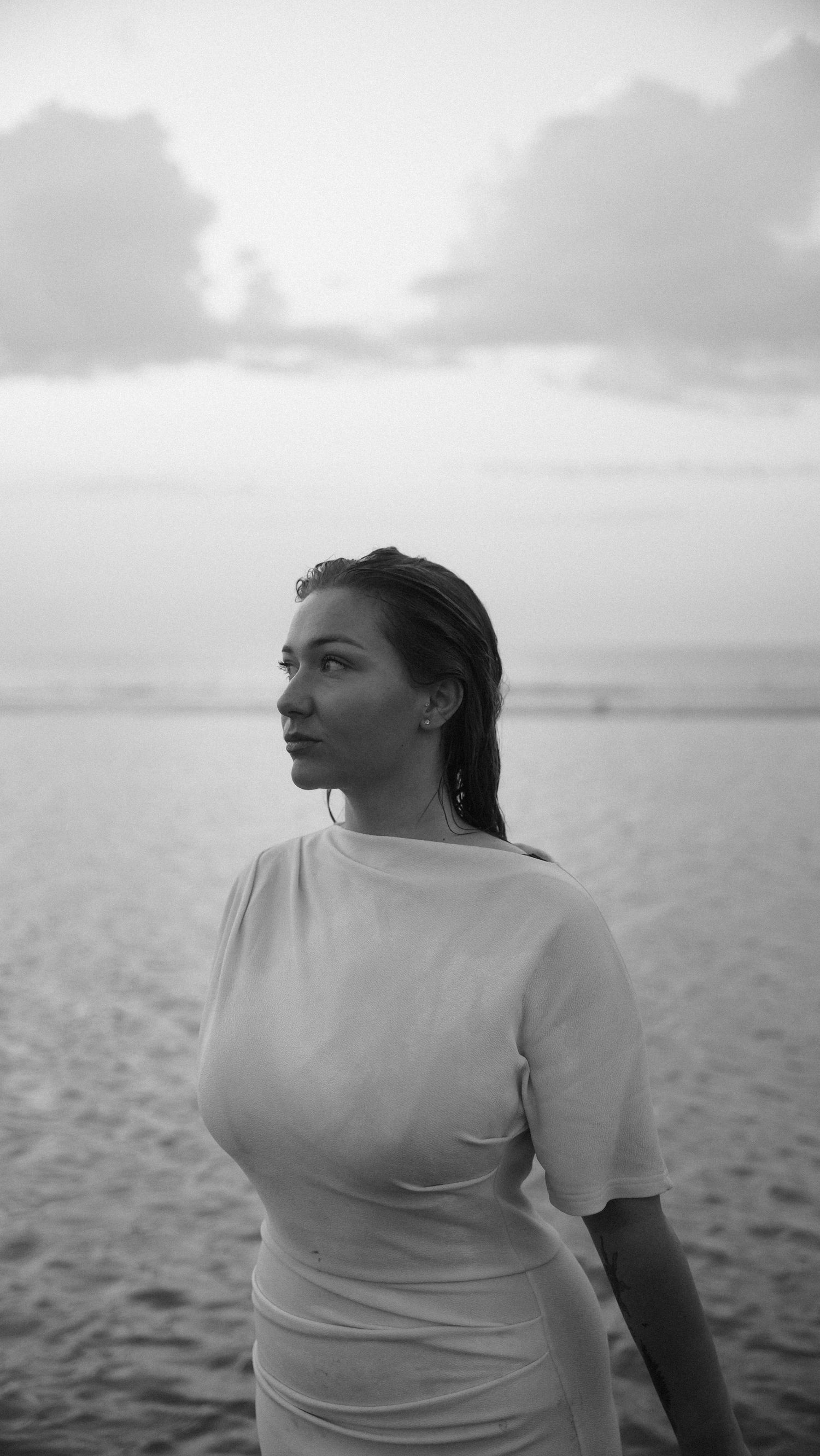 Black and white portrait of a woman in white dress standing by the sea at sunset, looking away. by Soft Sacred Studios