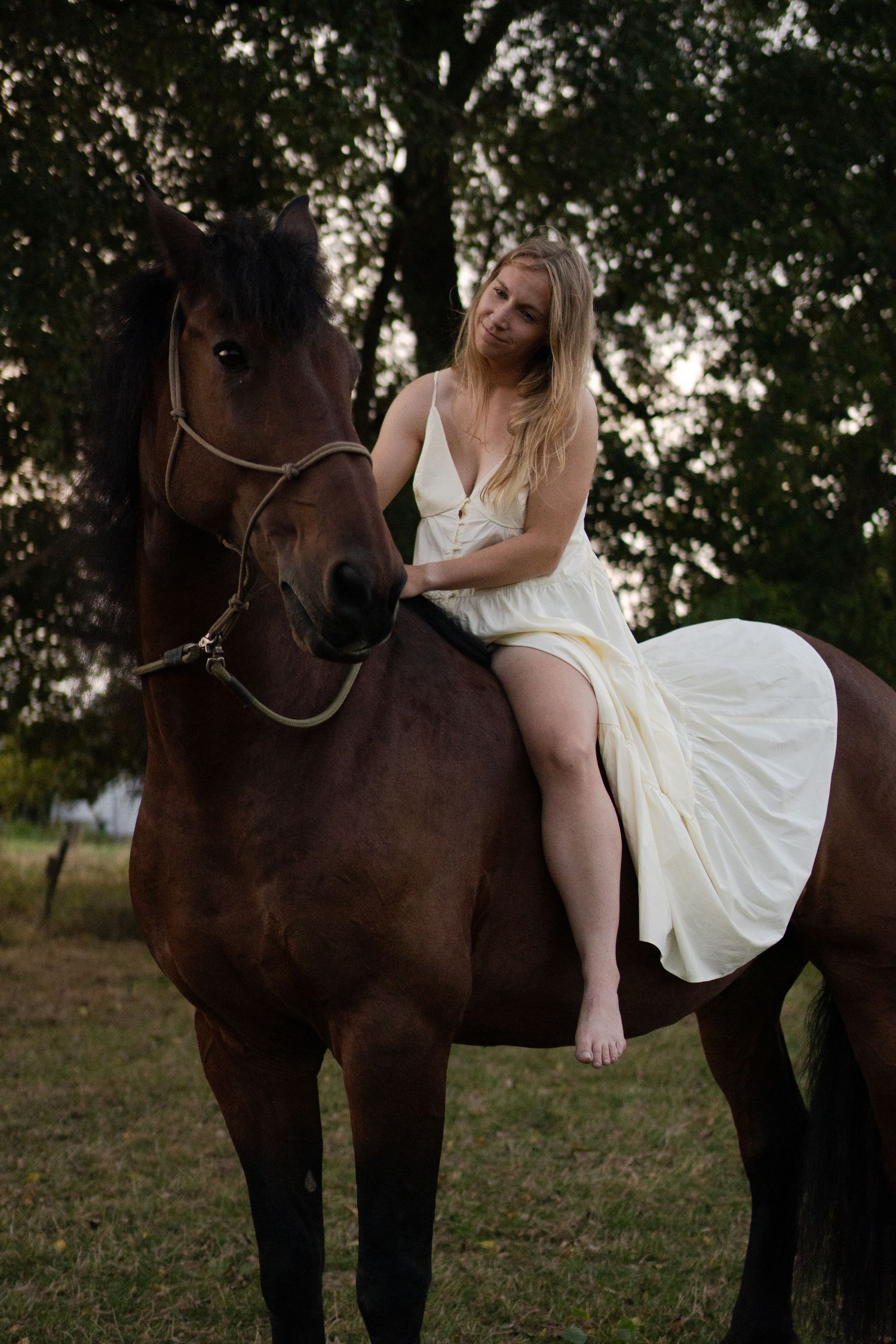 Smiling blonde woman in white dress riding bareback on a dark brown horse near trees at dusk. by Soft Sacred Studios