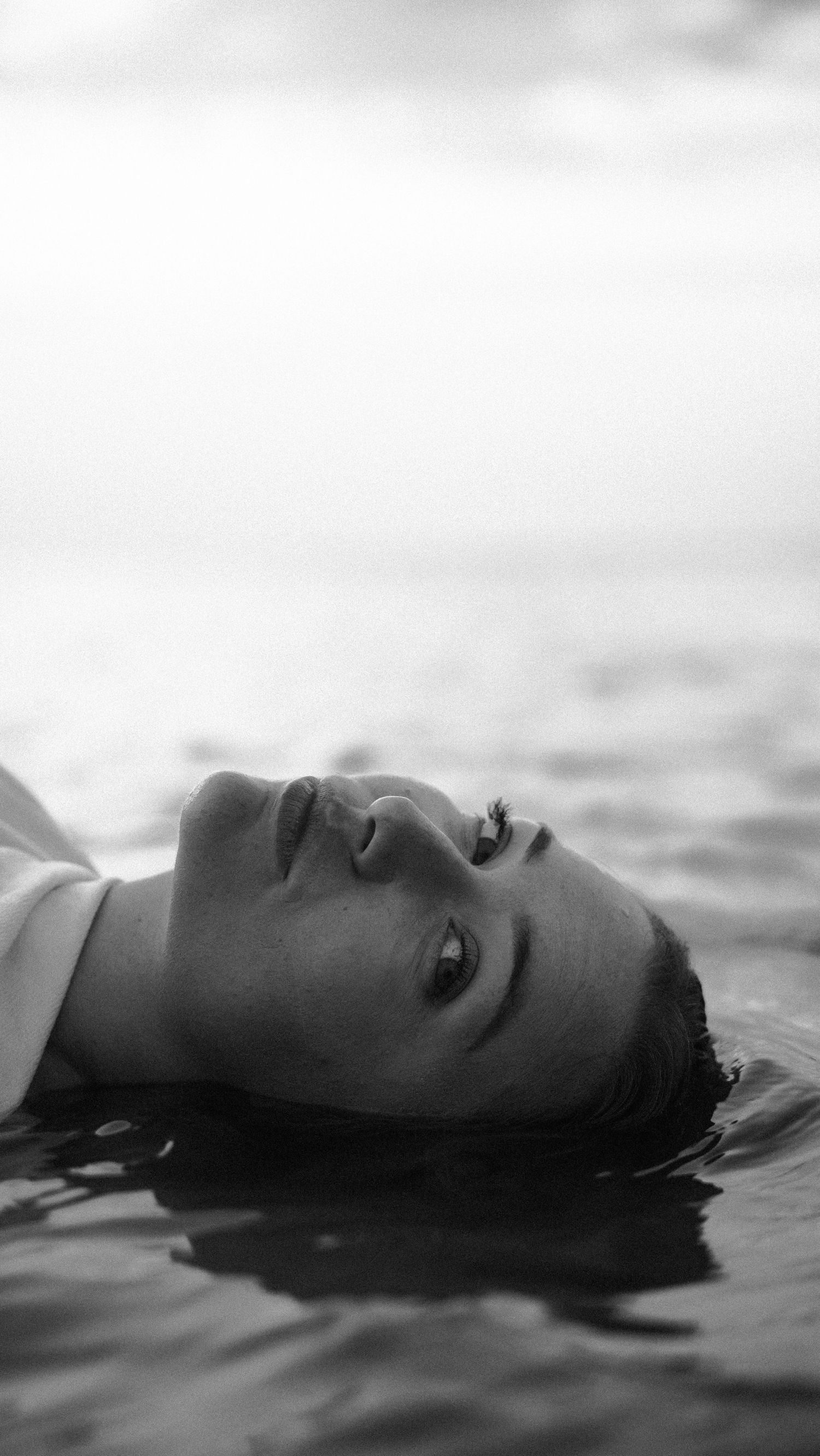 Close-up black and white portrait of woman floating in ocean water, serene expression. by Soft Sacred Studios
