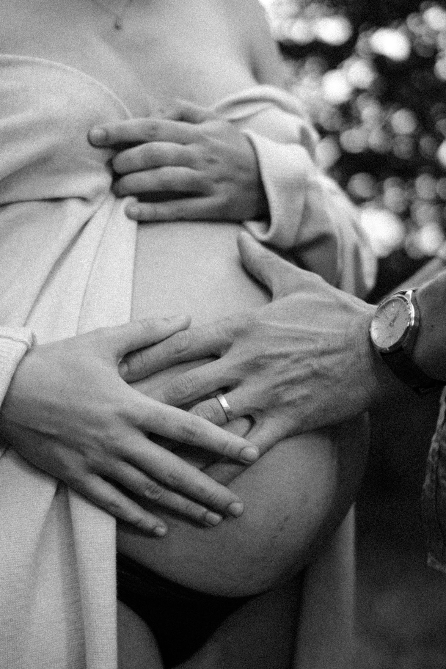 Black and white close-up of couple holding pregnant belly outdoors, man wearing wedding ring and watch. By Soft Sacred Studios.