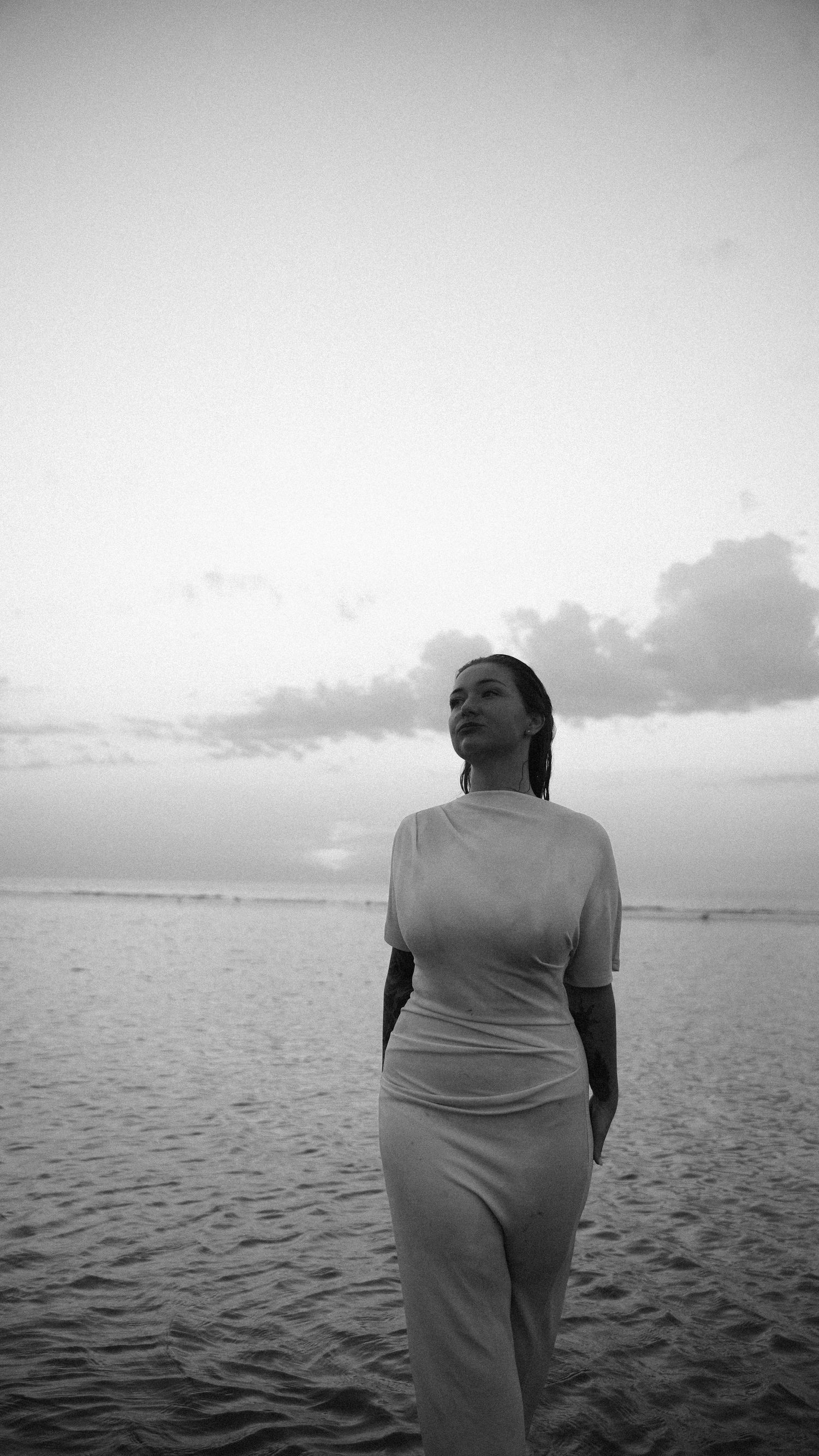 Woman in white dress standing on sandy beach at dusk, elegant black and white photo. by Soft Sacred Studios