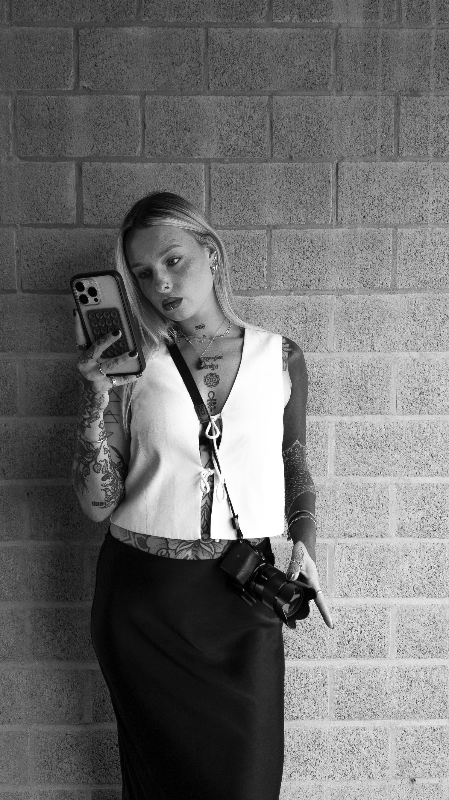 Tattooed blonde woman in white top and black skirt takes mirror selfie while holding camera against brick wall. By Soft Sacred Studios.