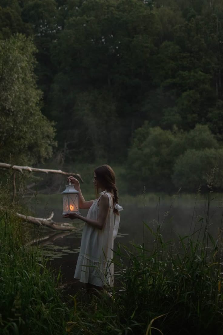 Woman in white dress holding a glowing lantern near a misty river at dusk. By Soft Sacred Studios.