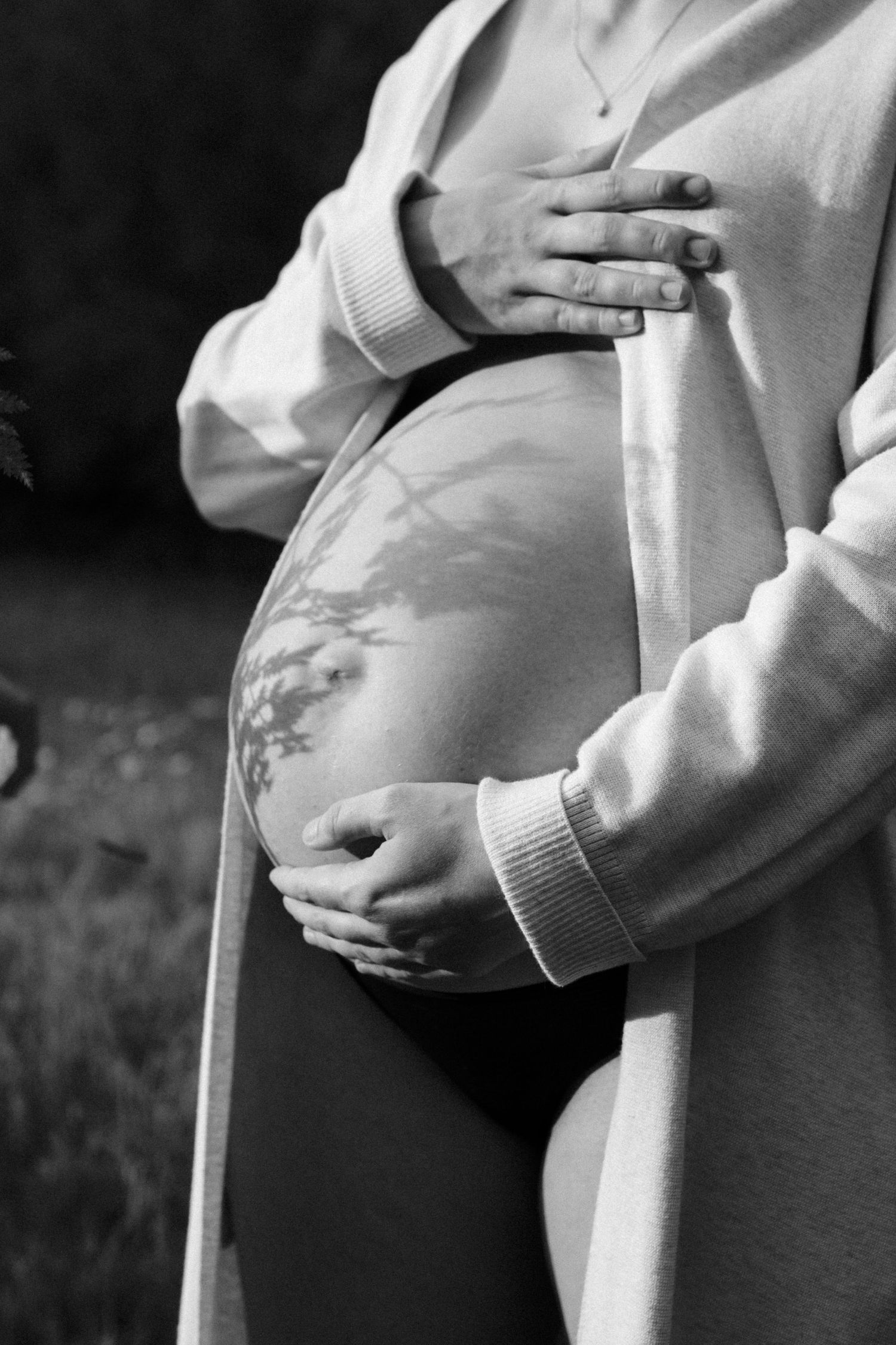 Black and white image of pregnant woman in cardigan cradling bare bump with delicate tree shadow on skin. by Soft Sacred Studios