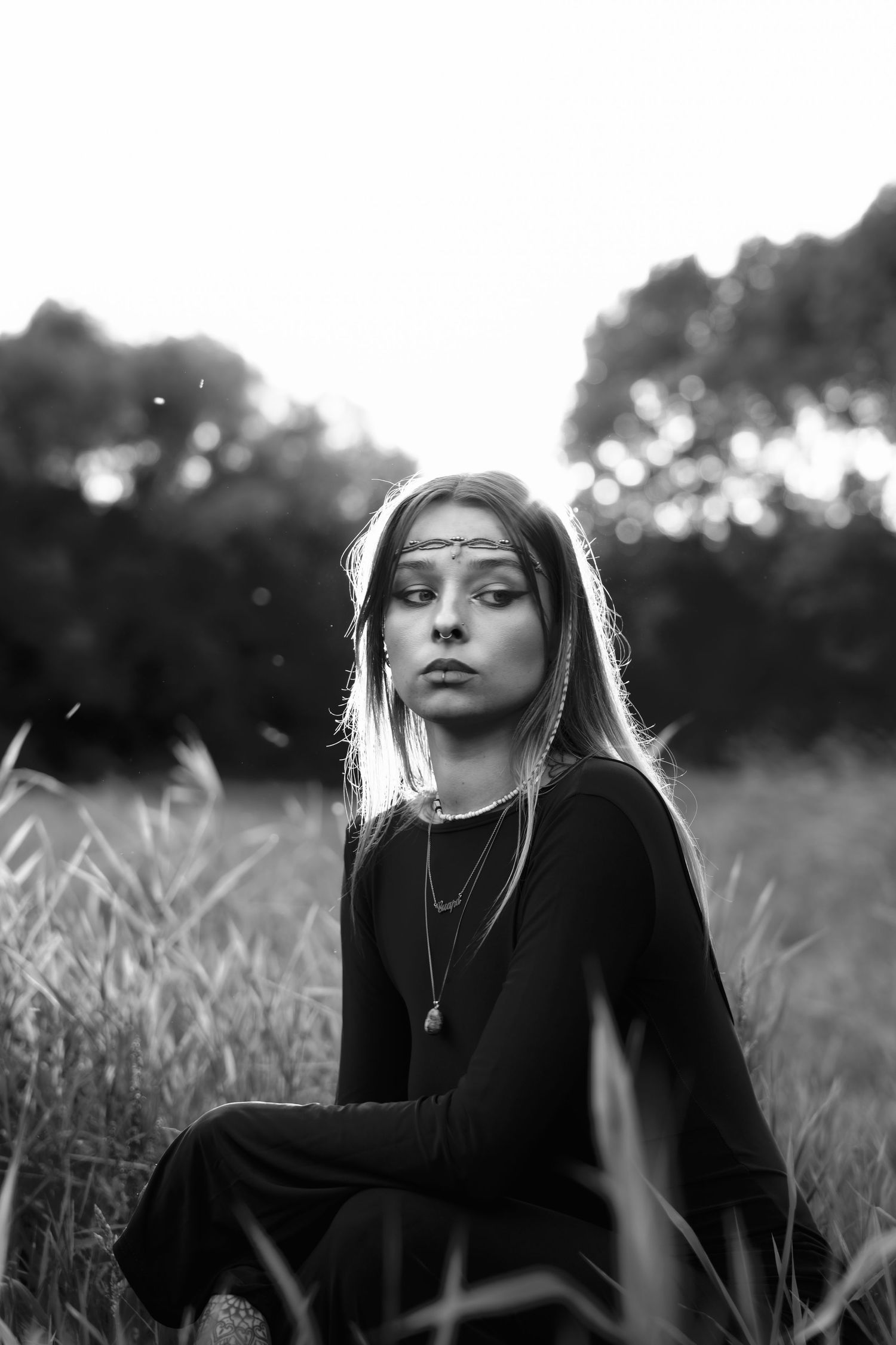 Black and white portrait of young woman in black dress sitting in tall grass field at dusk. By Soft Sacred Studios.