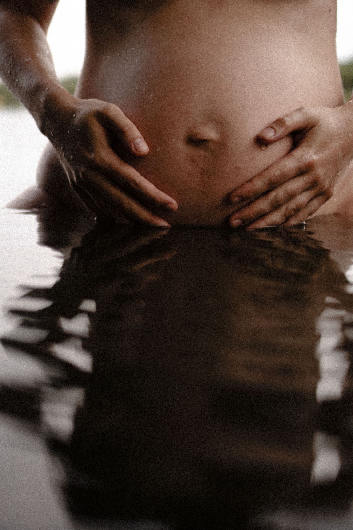 Close-up of pregnant belly with hands cradling bump reflected in still water surface. By Soft Sacred Studios.