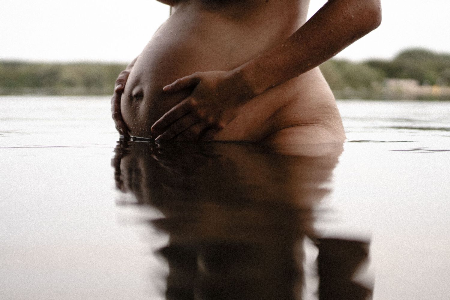 Artistic black and white maternity photo of pregnant belly being cradled by hands in still water. By Soft Sacred Studios.