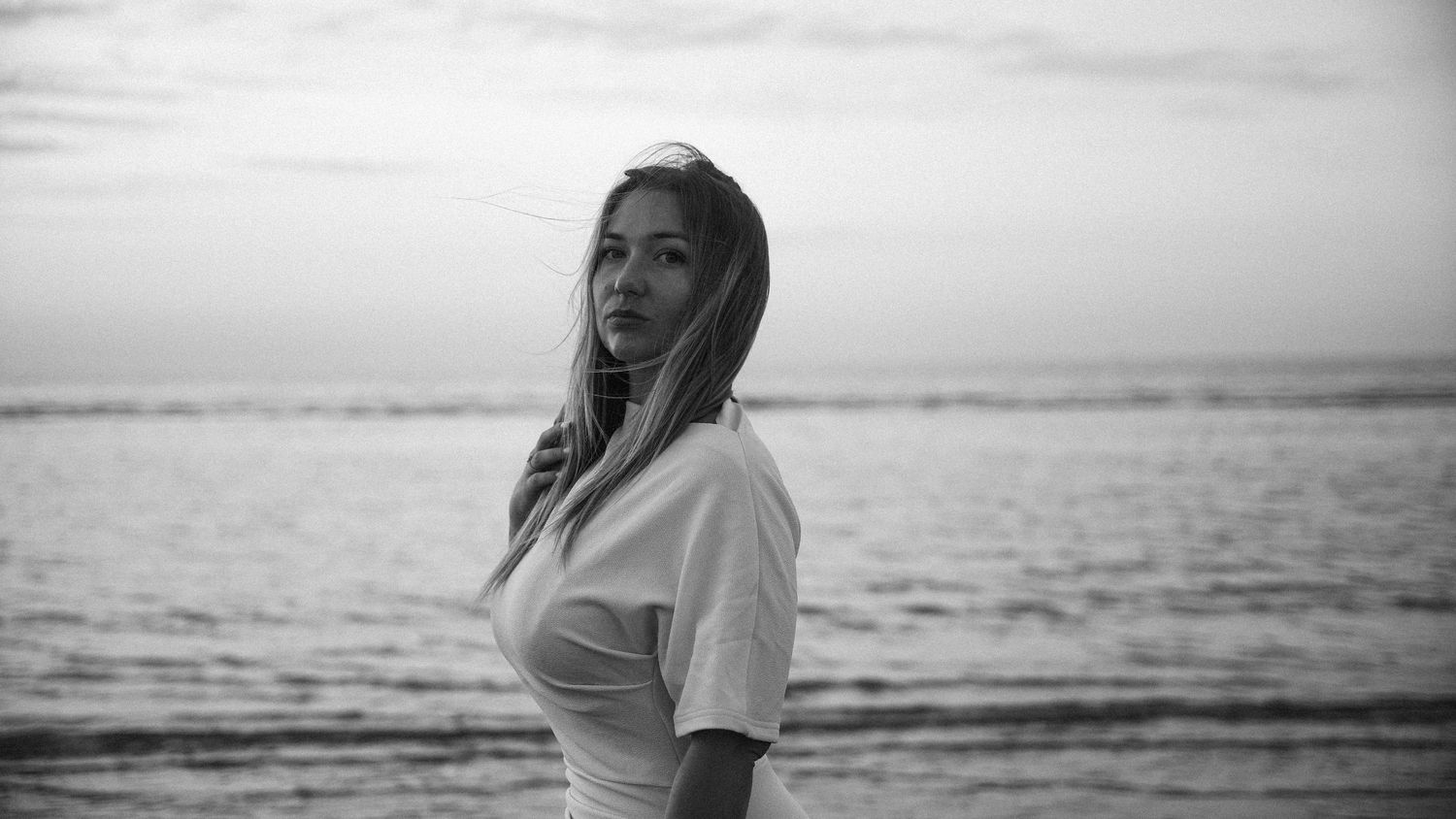 Young woman in white top posing on beach at sunset, black and white portrait photography. by Soft Sacred Studios