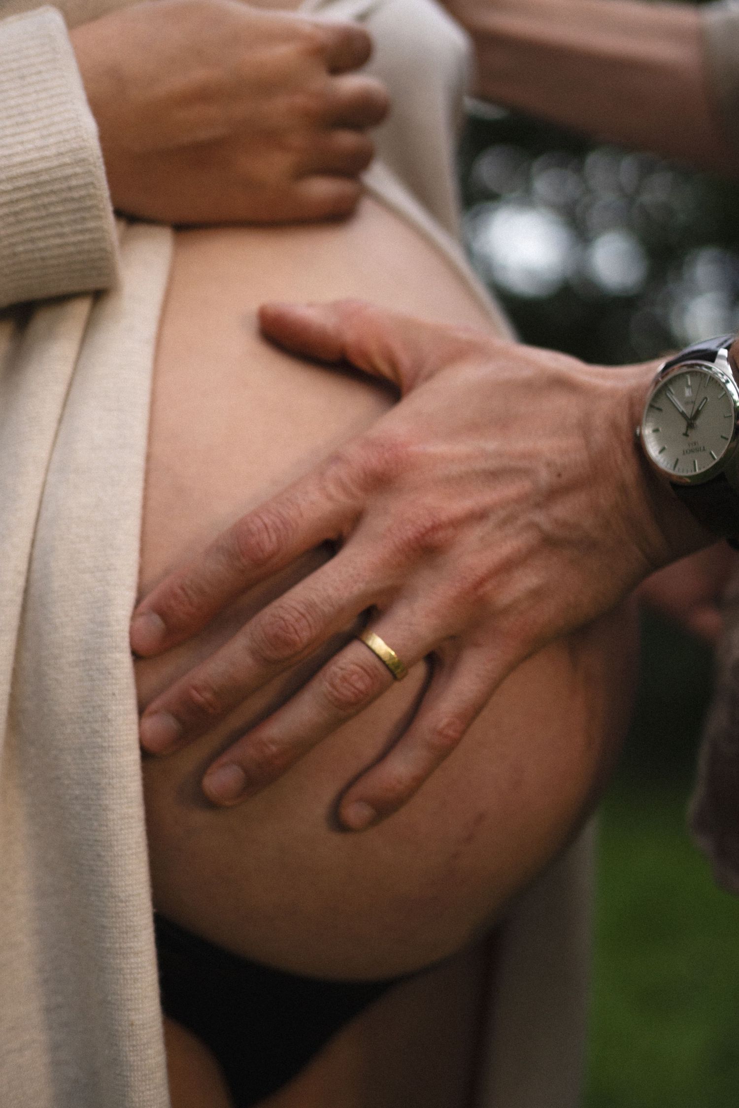 Close-up of partners hands with wedding ring gently holding pregnant womans bare baby bump. By Soft Sacred Studios.