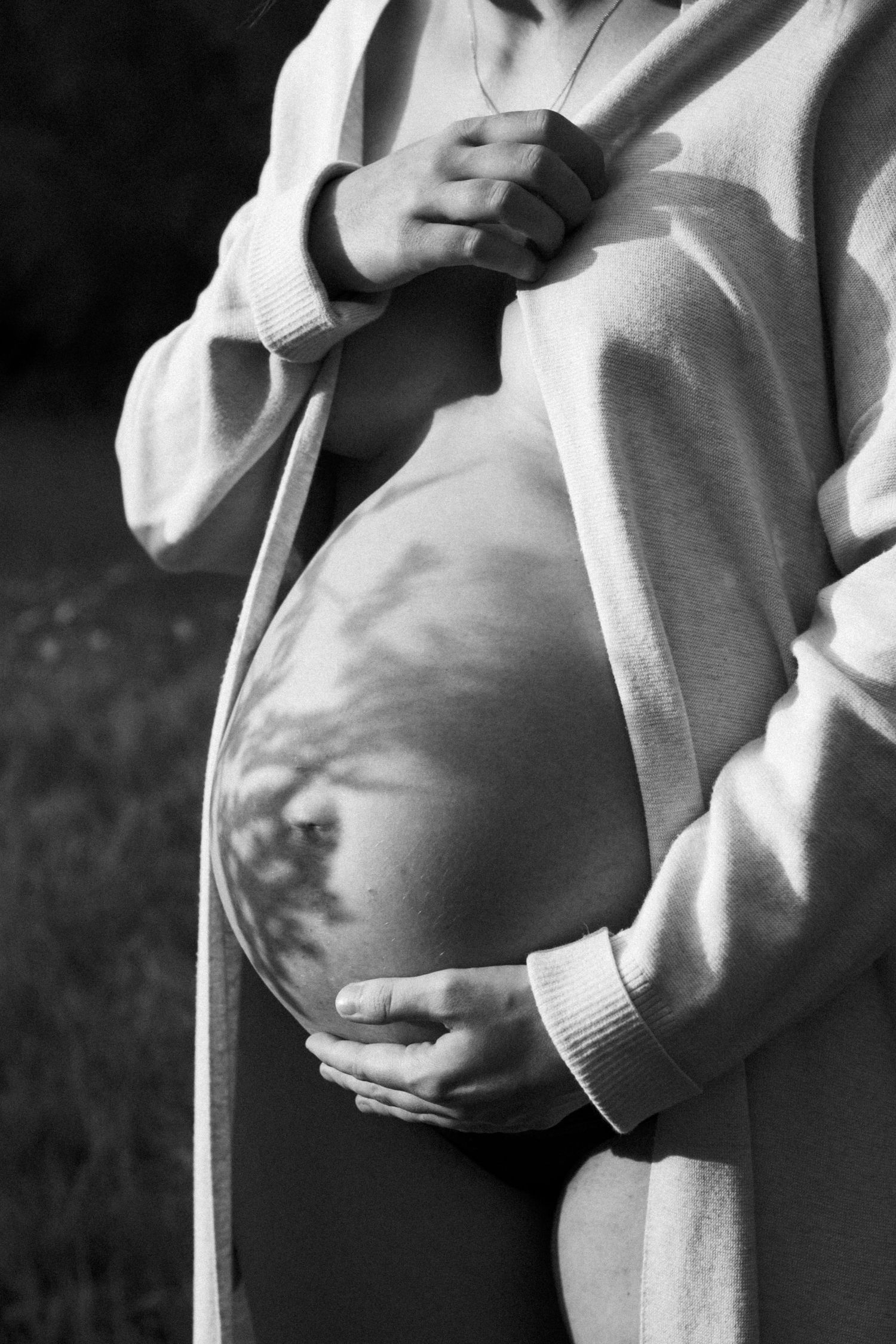 Black and white photo of pregnant woman in cozy cardigan cradling her baby bump outdoors. By Soft Sacred Studios.