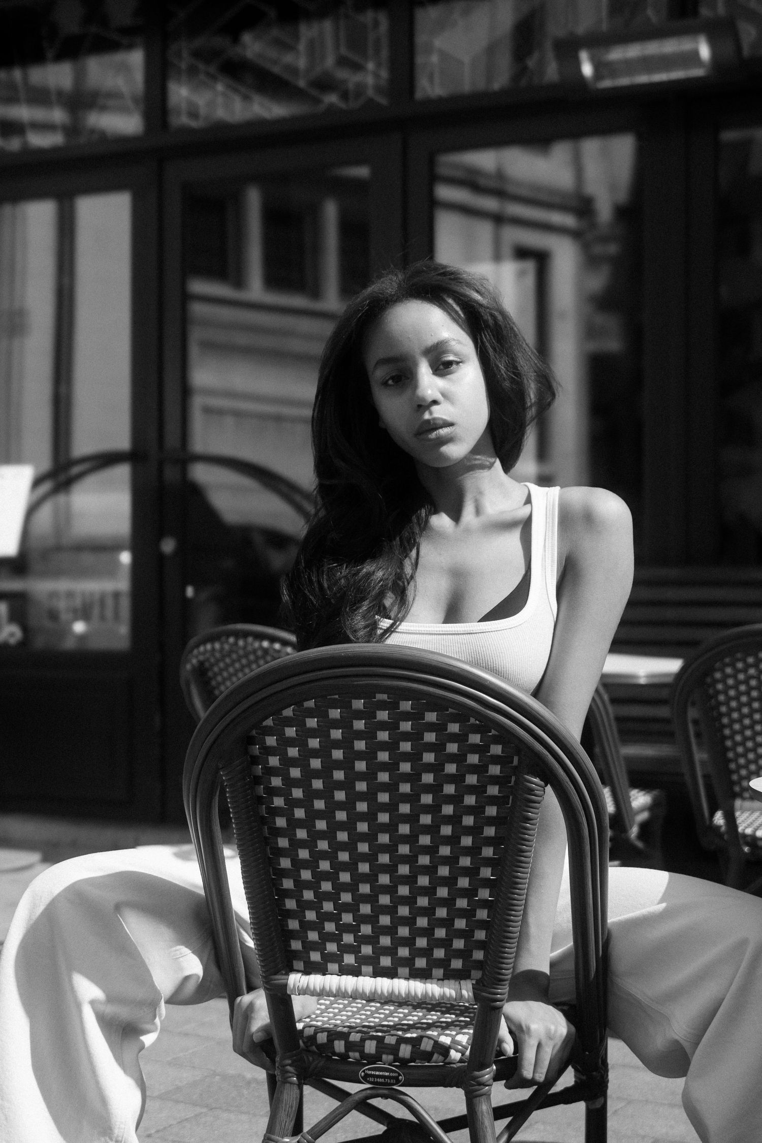 Black and white photo of young woman in tank top posing at outdoor cafe chair. By Soft Sacred Studios.