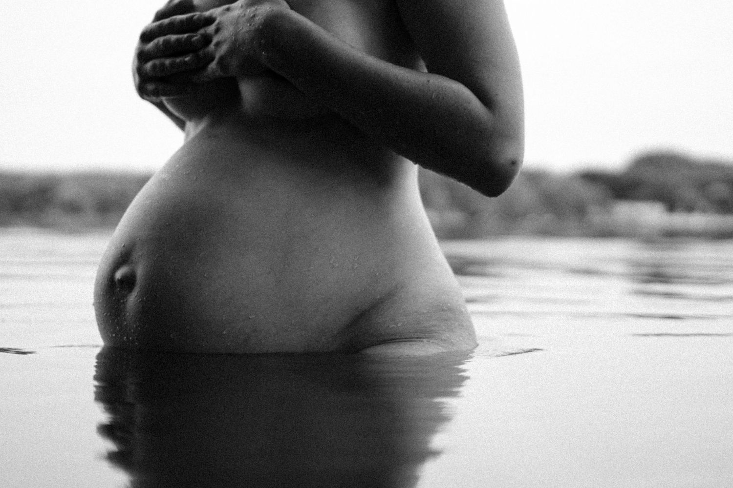 Black and white side profile of nude pregnant woman standing in calm water with arms crossed. By Soft Sacred Studios.