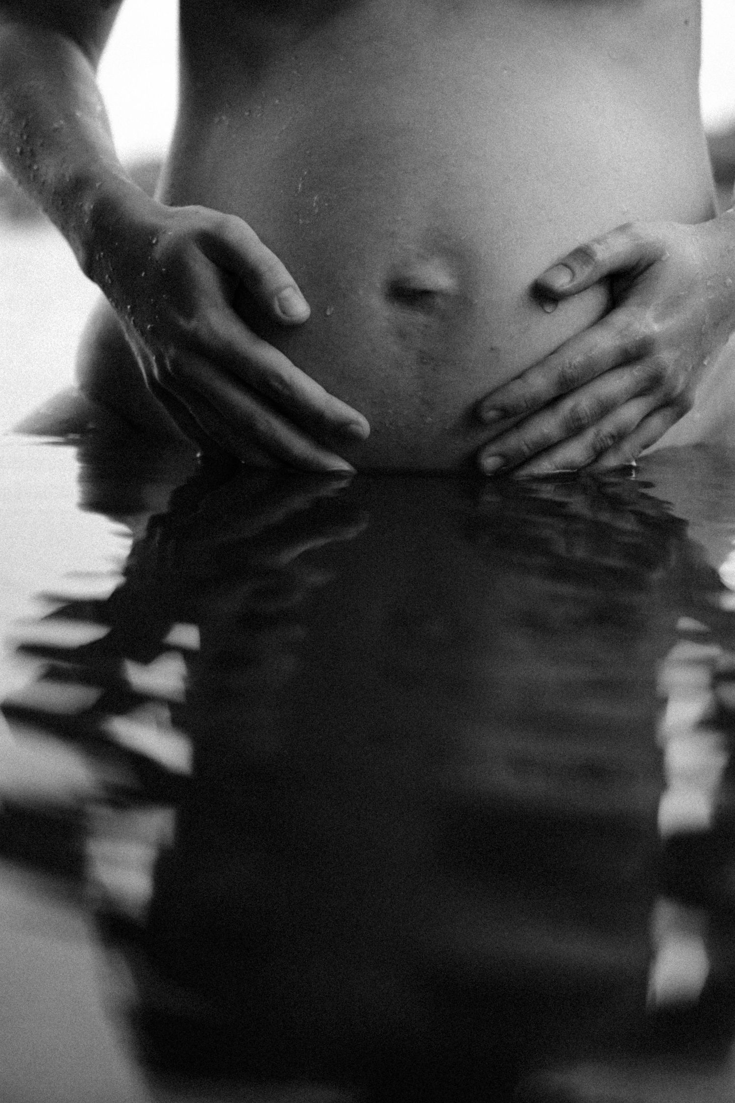 Black and white photo of pregnant woman standing in water with hands cradling baby bump. By Soft Sacred Studios.