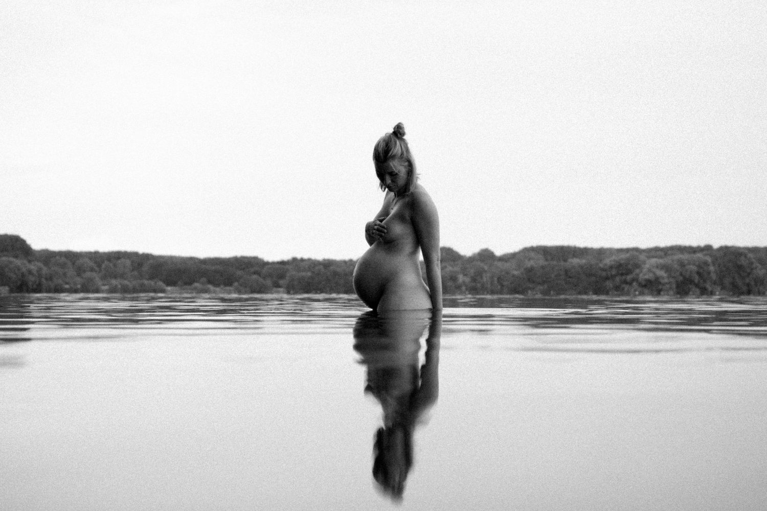 Black and white silhouette of pregnant woman standing in calm reflective lake water outdoors. By Soft Sacred Studios.