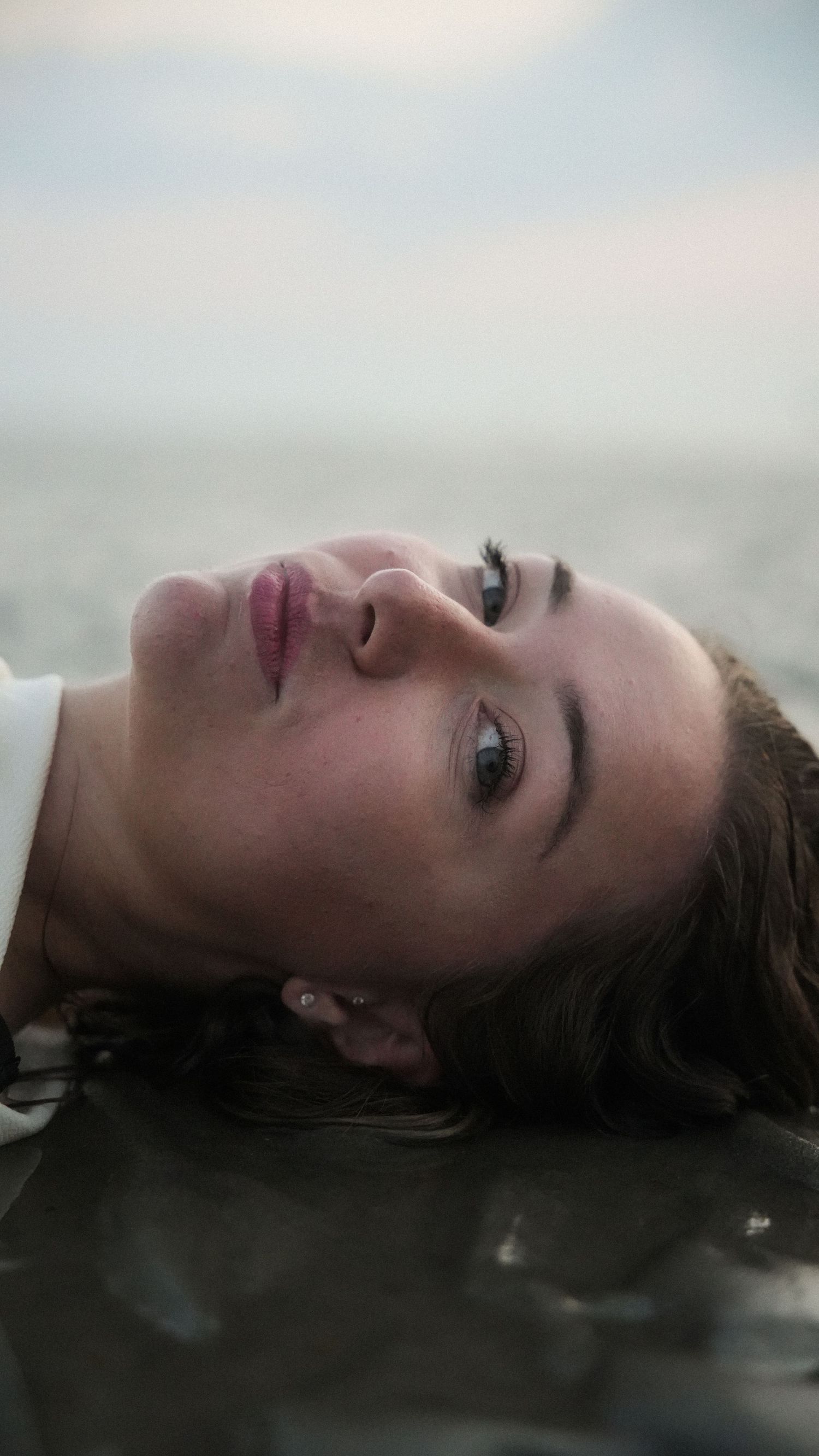 Close-up portrait of a woman lying in shallow ocean water, looking up with striking blue eyes. by Soft Sacred Studios