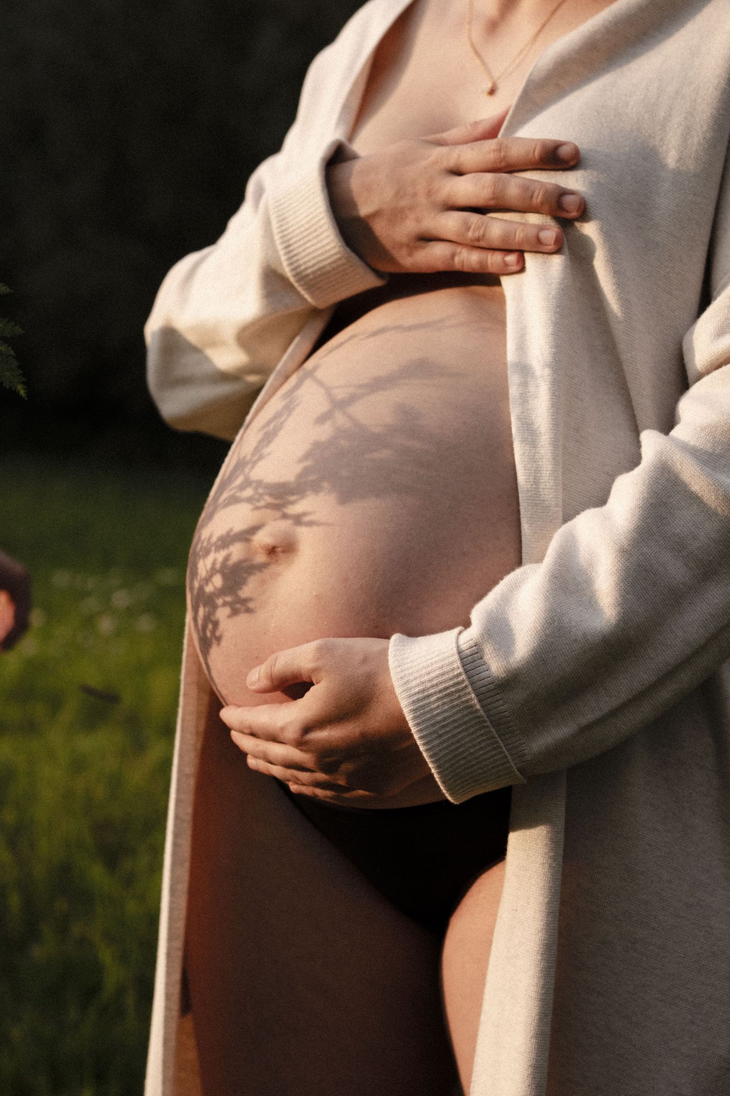 Close-up of pregnant belly with tree branch shadow pattern, hands cradling bump in open cream cardigan. by Soft Sacred Studios