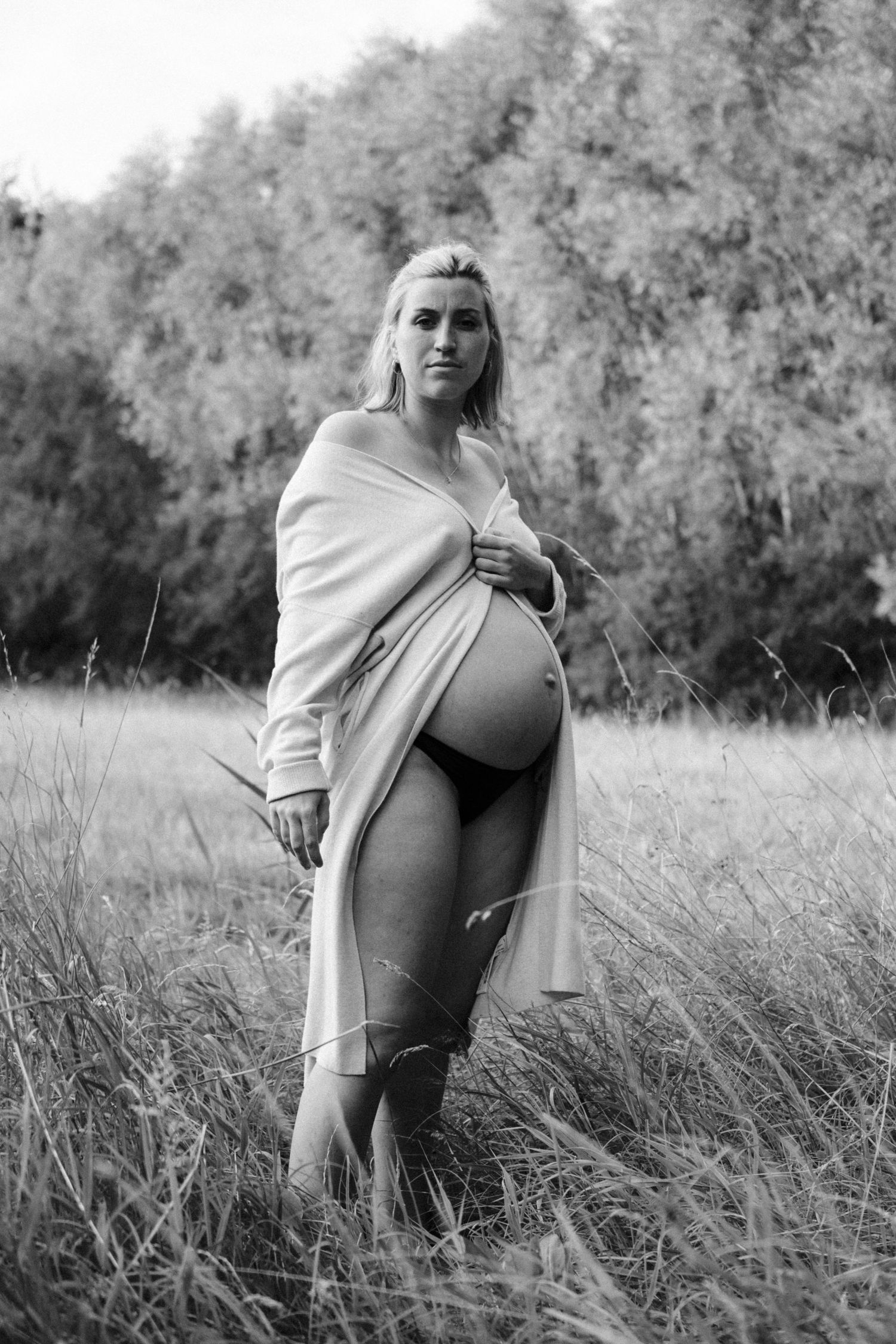Black and white portrait of pregnant blonde woman in open cardigan standing in tall grass field outdoors. By Soft Sacred Studios.