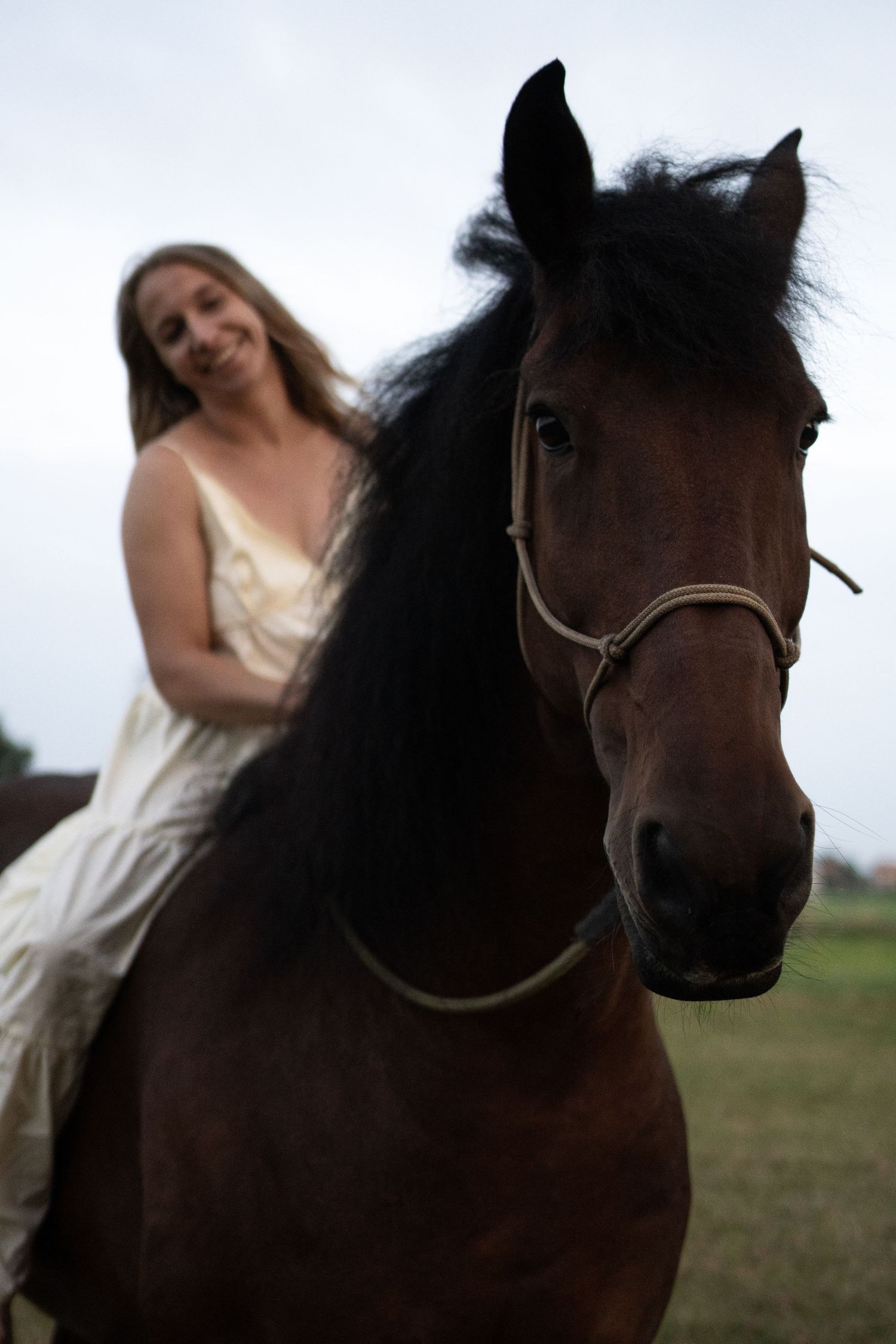 Smiling blonde woman in white dress riding a dark brown horse with flowing black mane. by Soft Sacred Studios
