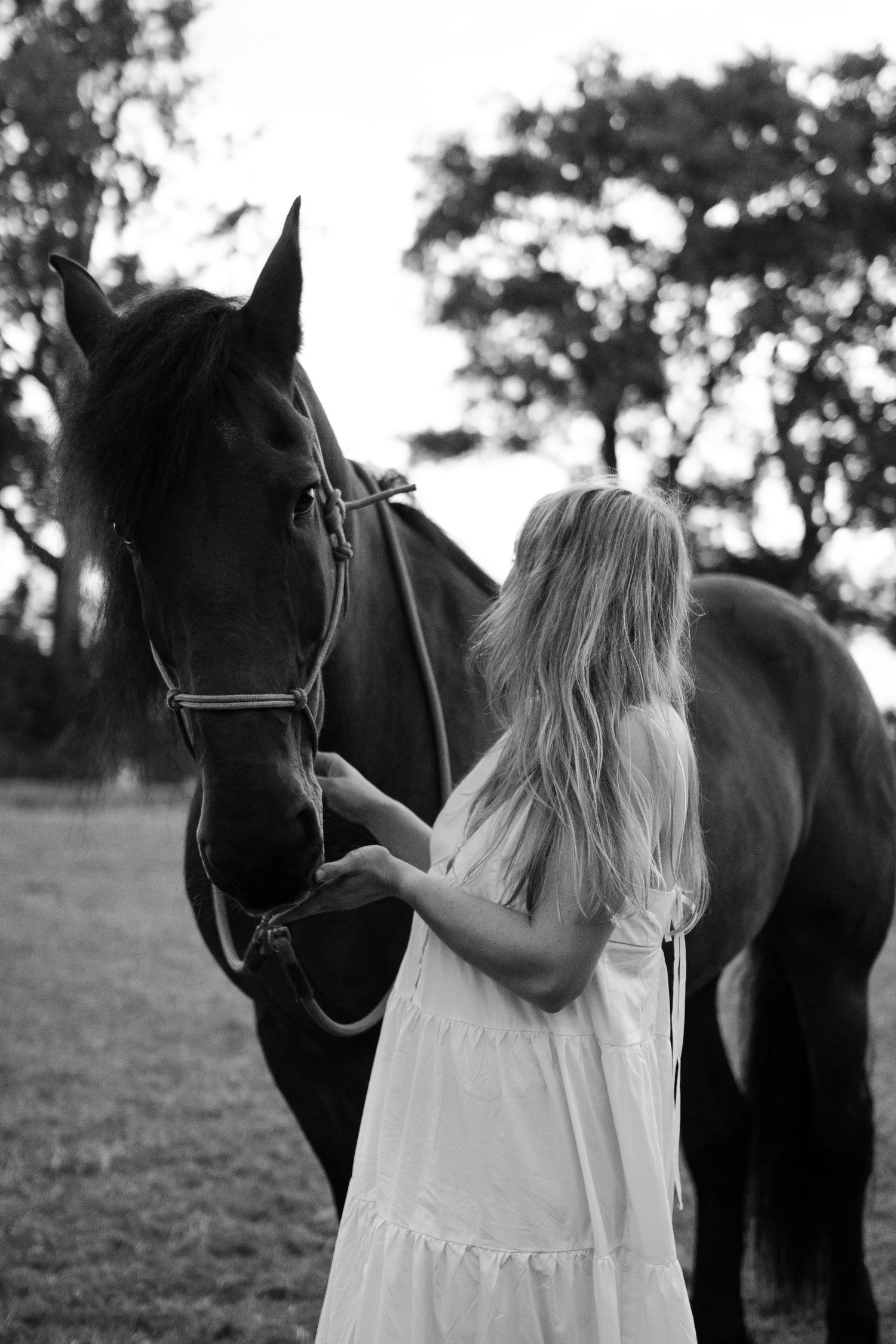 Black and white photo of blonde woman in white dress standing with a dark horse in a field. by Soft Sacred Studios