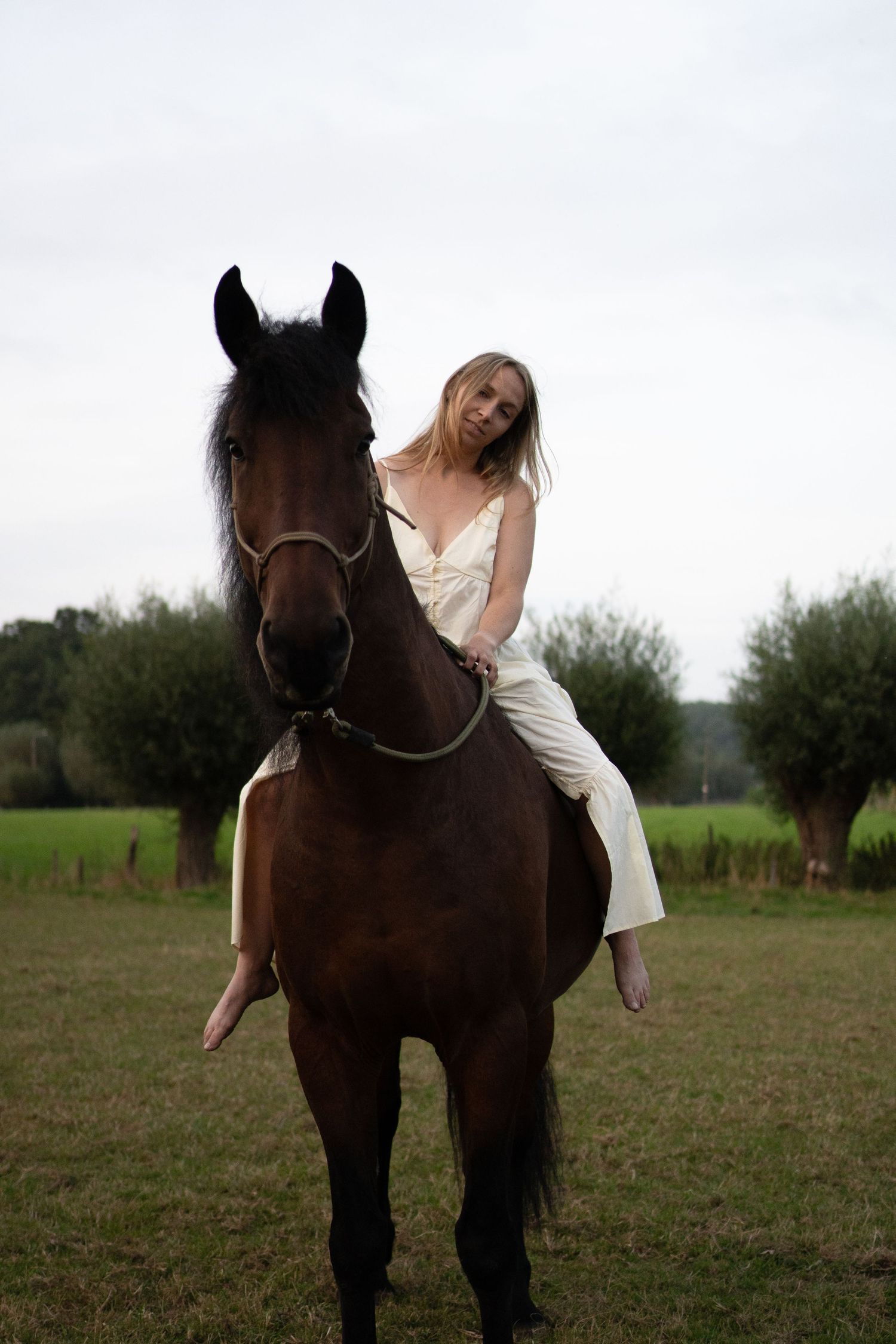 Blonde woman in white outfit lying forward on a dark brown horse in a green pasture. by Soft Sacred Studios