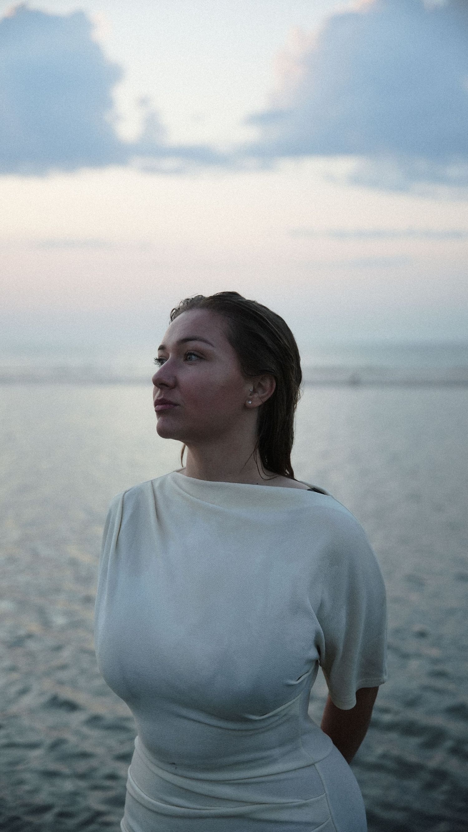 Woman in white dress standing by the ocean at dusk, gazing into the distance with a serene expression. by Soft Sacred Studios