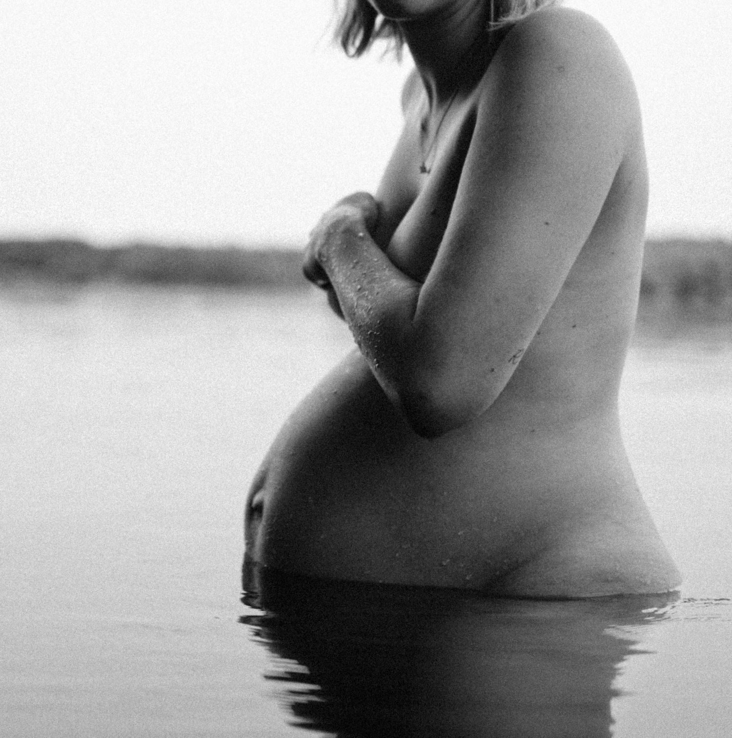 Black and white side profile of nude pregnant woman standing in calm water, arms folded across chest. By Soft Sacred Studios.